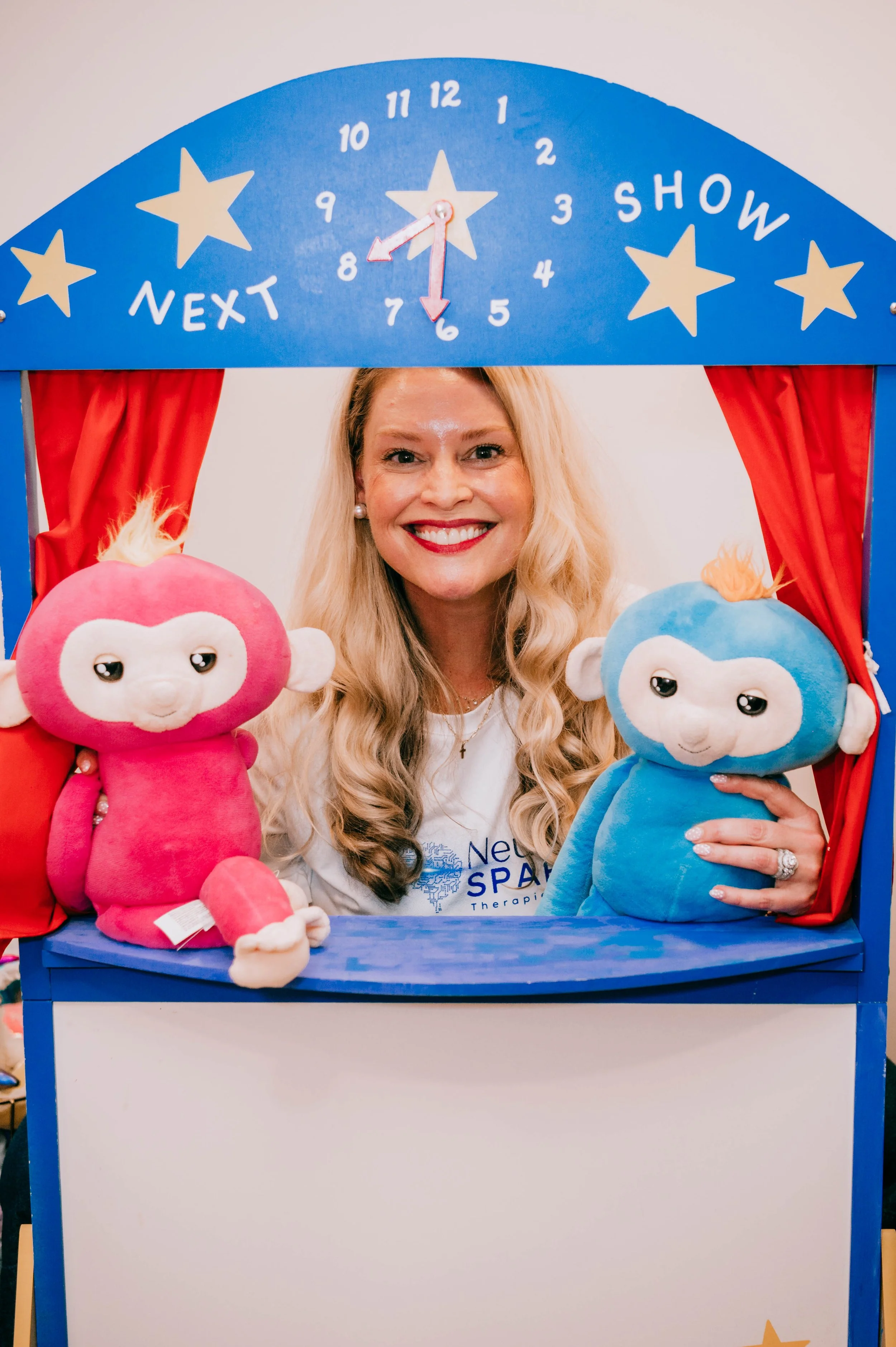 A woman with long blonde hair smiling, sitting with a pink pig stuffed animal, wearing a black t-shirt and a watch, with a decorated green wall and black acoustic panels behind her.