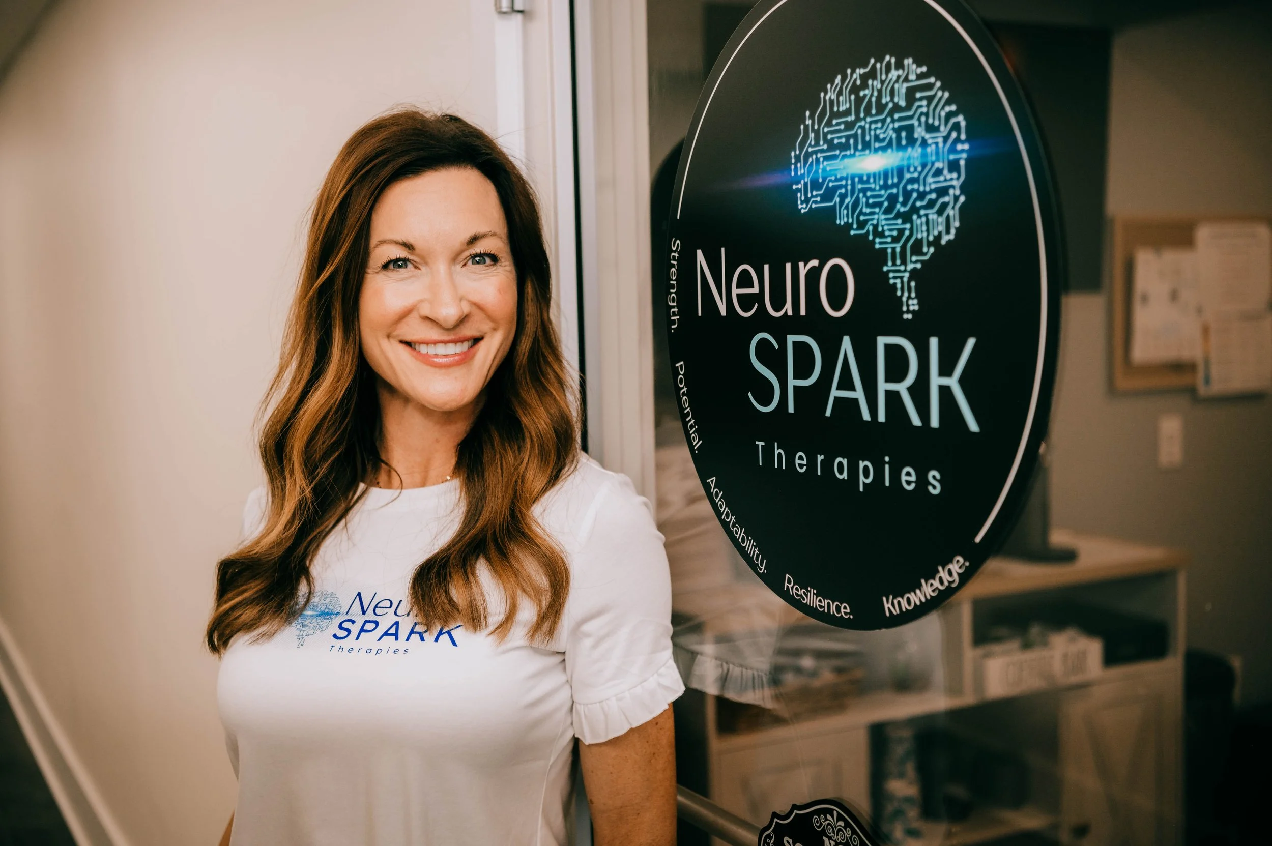 Woman smiling in front of a sign that says 'Neuro SPARK Therapies' with a brain graphic.