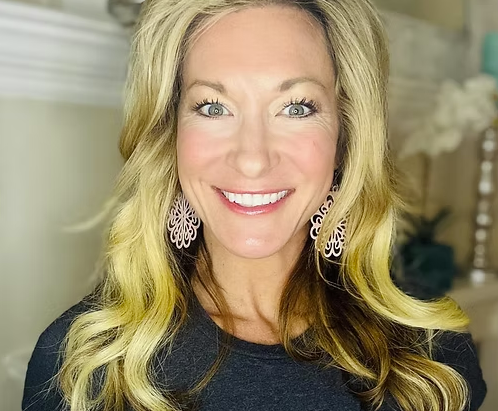 A woman with long blonde hair, smiling, wearing purple earrings and a dark top, in an indoor setting.