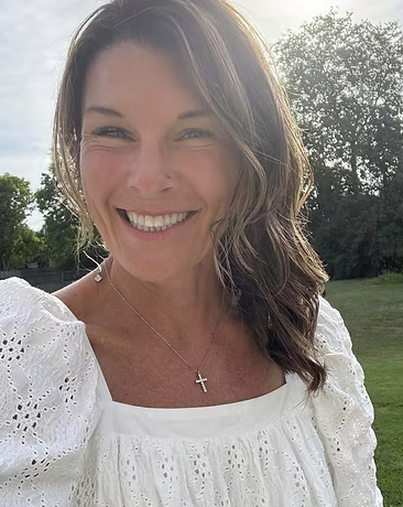 A woman with brown hair smiling, wearing a white blouse with eyelet details and a cross necklace, outdoors on a sunny day with trees in the background.