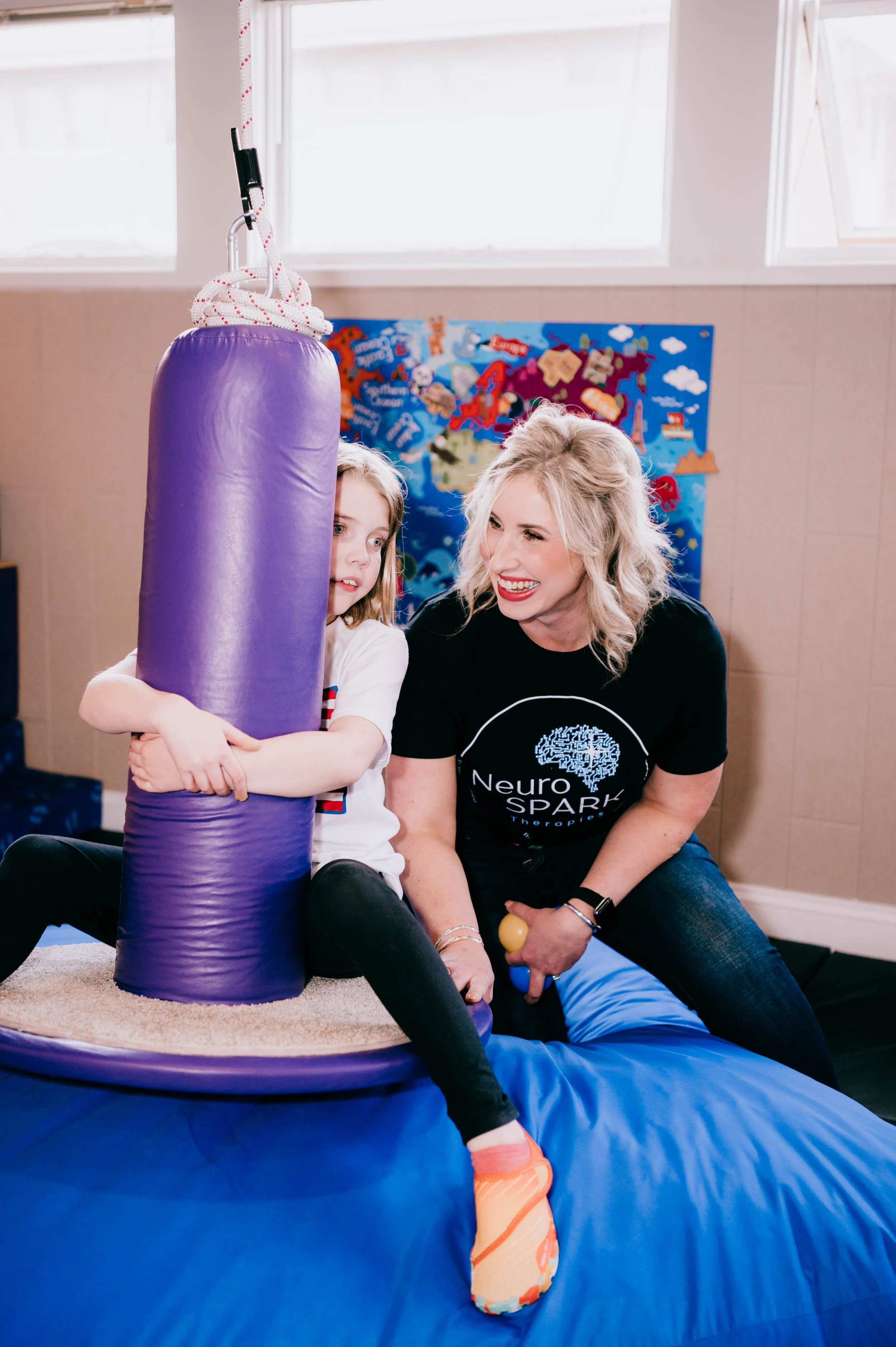 A woman with blonde hair and a young girl hang out in a sensory room, playing with a purple crash pad and a yellow ball, against a backdrop of colorful decorations and a world map poster.