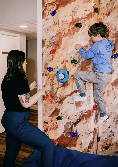A young boy climbing a rock wall with colorful holds, supervised by an adult woman in an indoor play area.