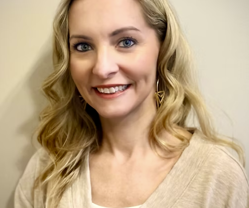 A young woman with blonde curly hair, blue eyes, and wearing gold earrings smiling in front of a plain beige wall.