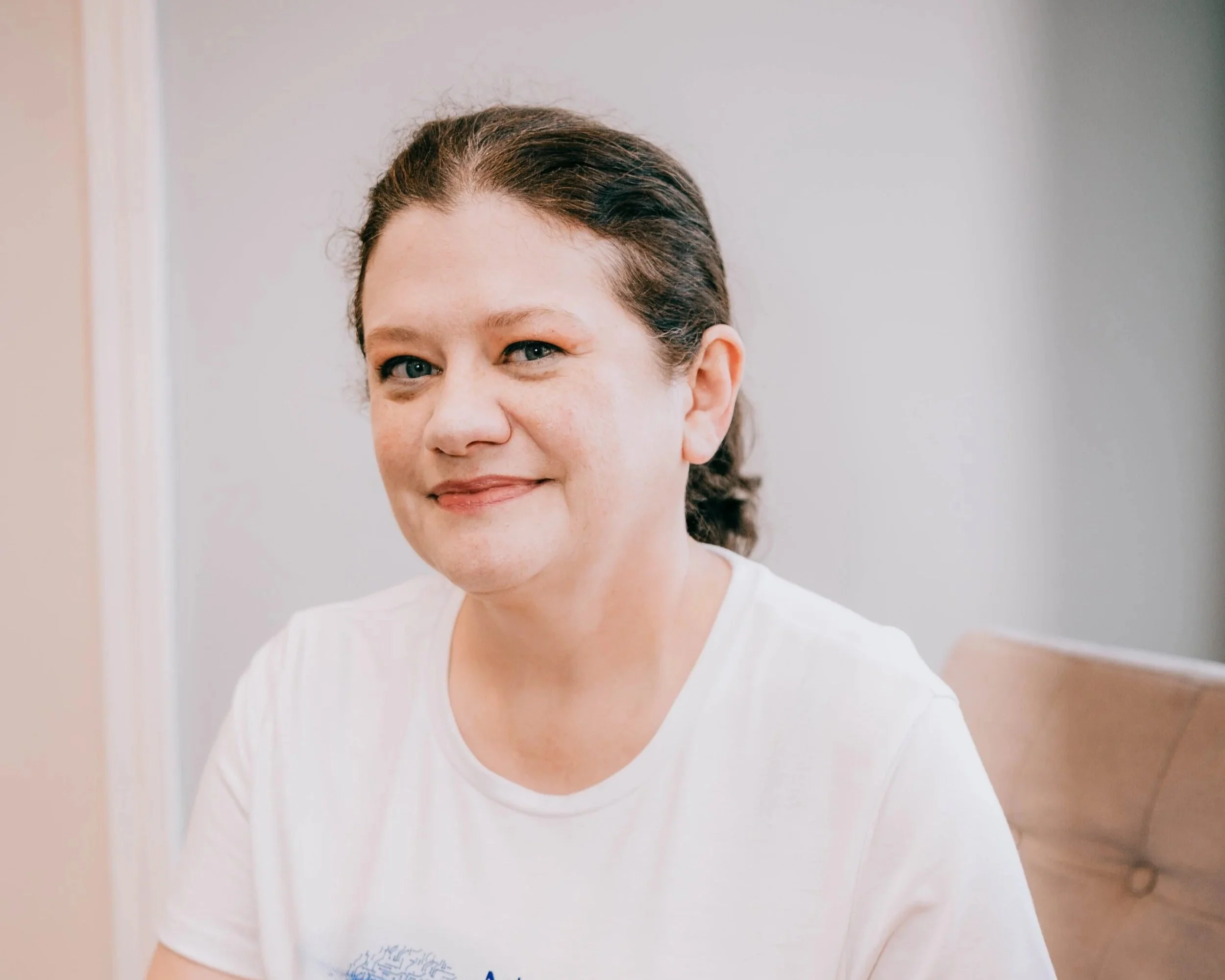 Close-up of a woman with dark brown hair, blue eyes, light skin, wearing a colorful patterned top, smiling, against a plain gray background.