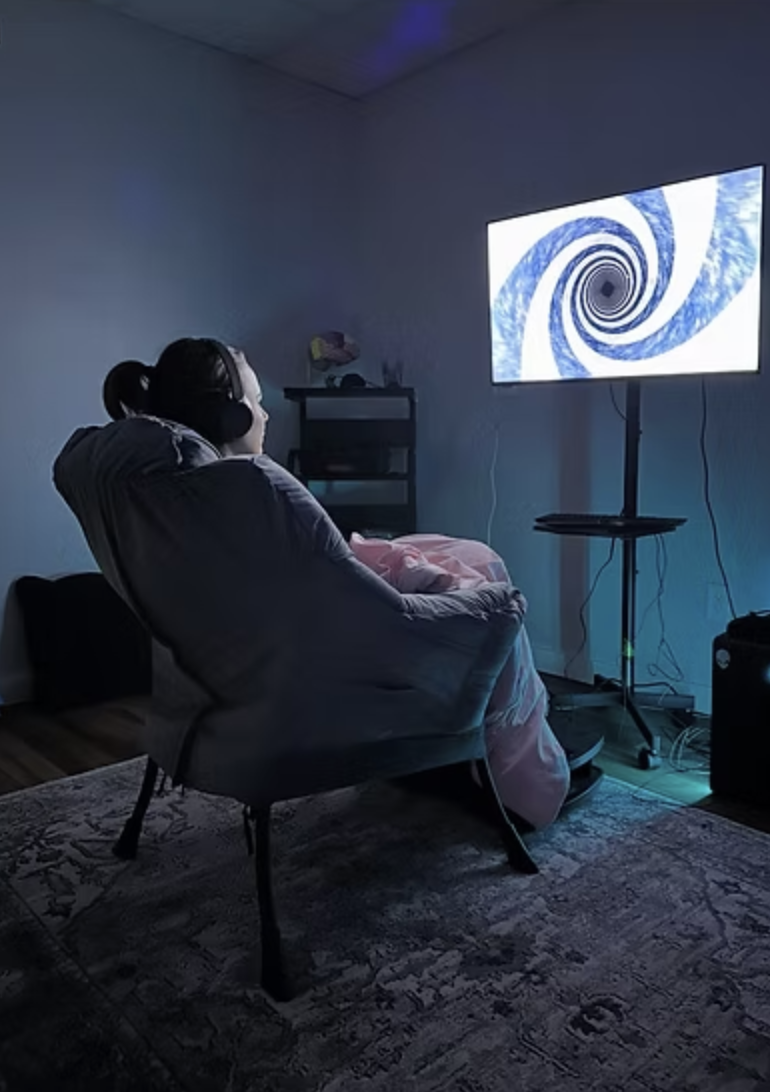 A person sitting in a reclined chair watching a television screen displaying a swirling hypnotic pattern in a dimly lit room
