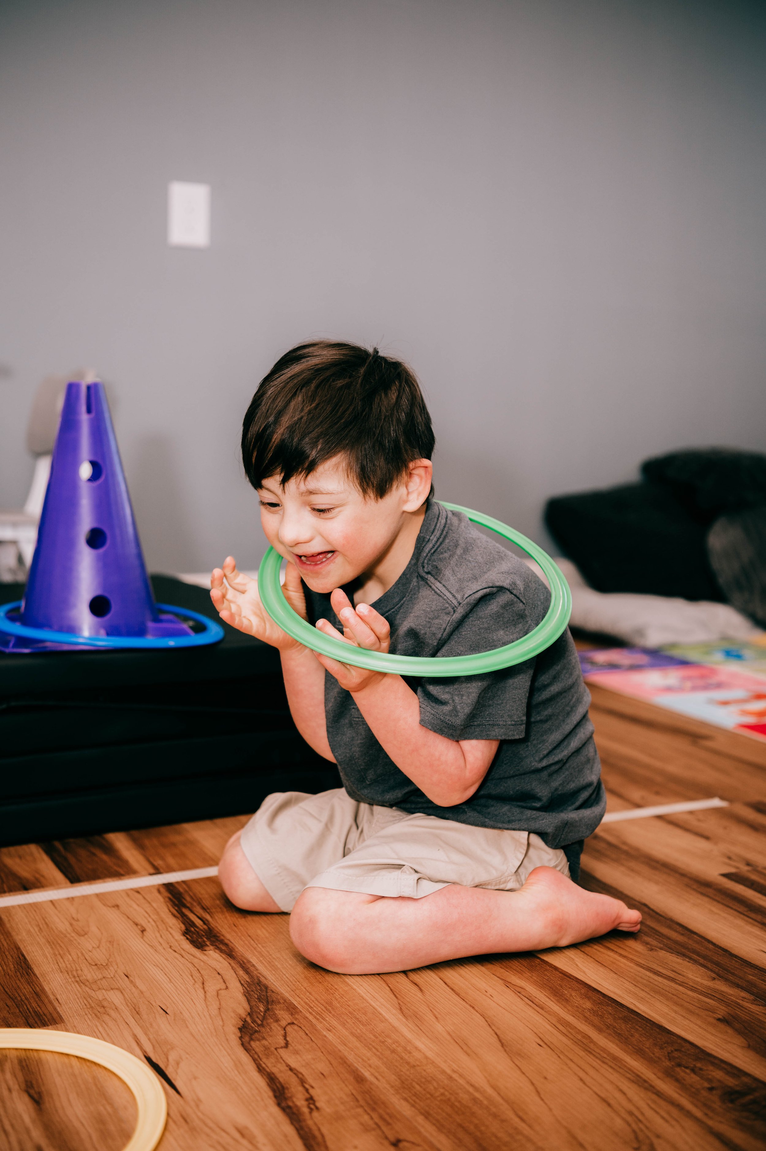 A young boy kneeling on a hardwood floor, smiling and playing with a green hoop around his neck, with a purple cone-shaped object in front of him.