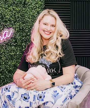 A woman with long blonde hair smiling, sitting with a pink pig stuffed animal, wearing a black t-shirt and a watch, with a decorated green wall and black acoustic panels behind her.