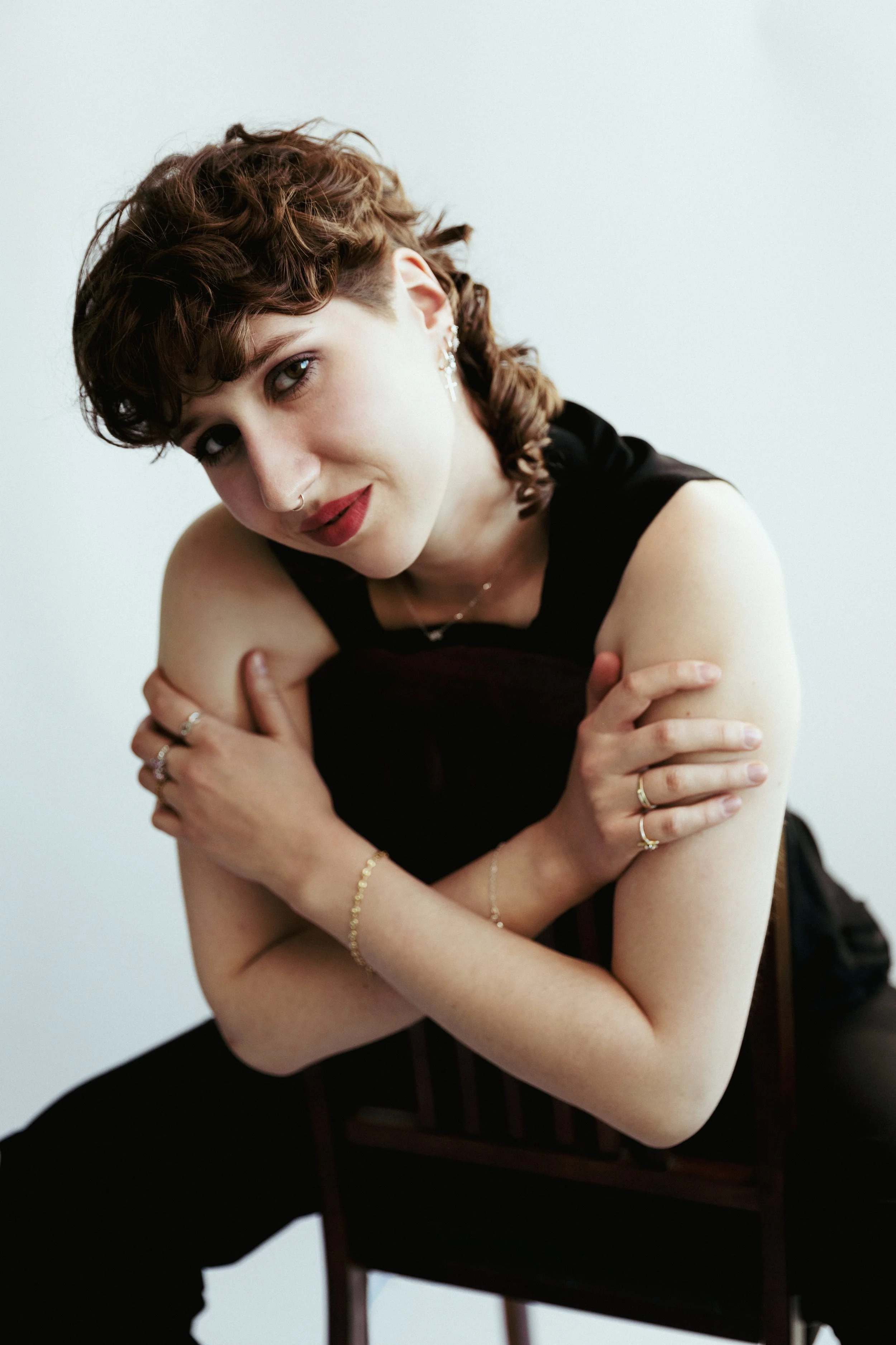 Junge Frau mit kurzen, braunen, lockigen Haaren und roten Lippen sitzt auf einem Stuhl, umarmt sich selbst, trägt schwarze Kleidung, Silberschmuck und schaut in die Kamera. Weißer Hintergrund.