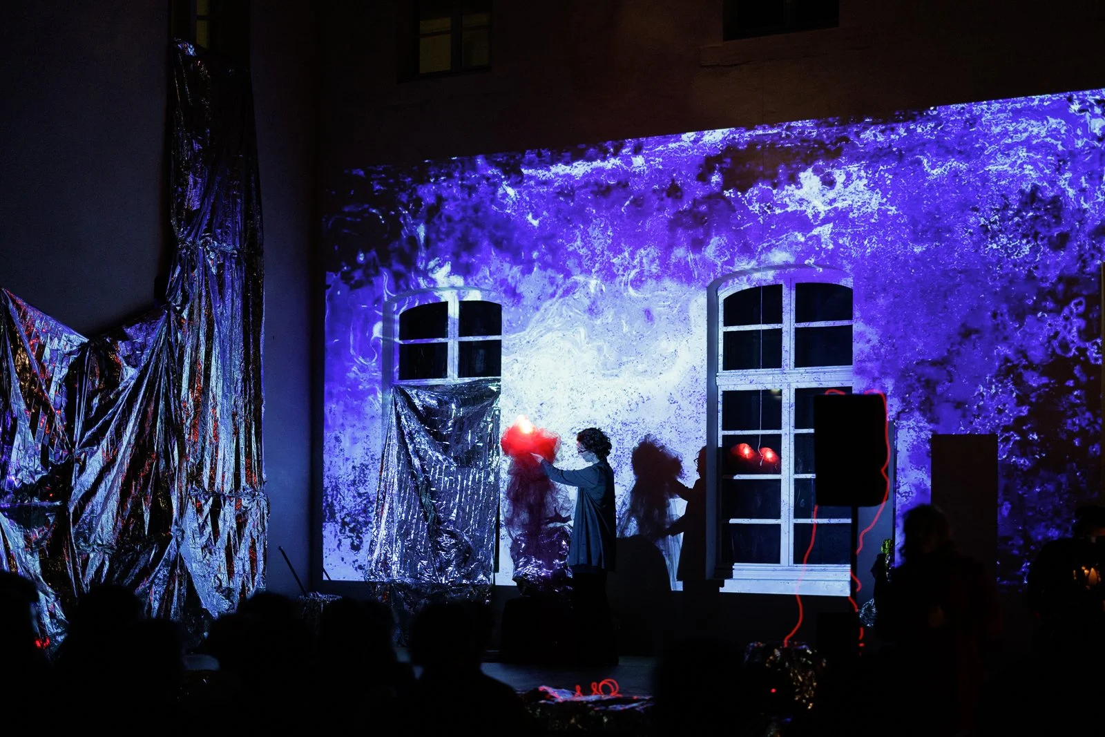 Eine dunkle Bühne mit einer abstrakten, leuchtend blauen und lila projizierten Wand hinter einem Fenster. Ein Mensch mit dunkler Kleidung hält eine leuchtende rote Kugel oder Lampe. Es gibt metallisch schimmernde Dekorationen und Schatten von anderen Personen im Publikum.