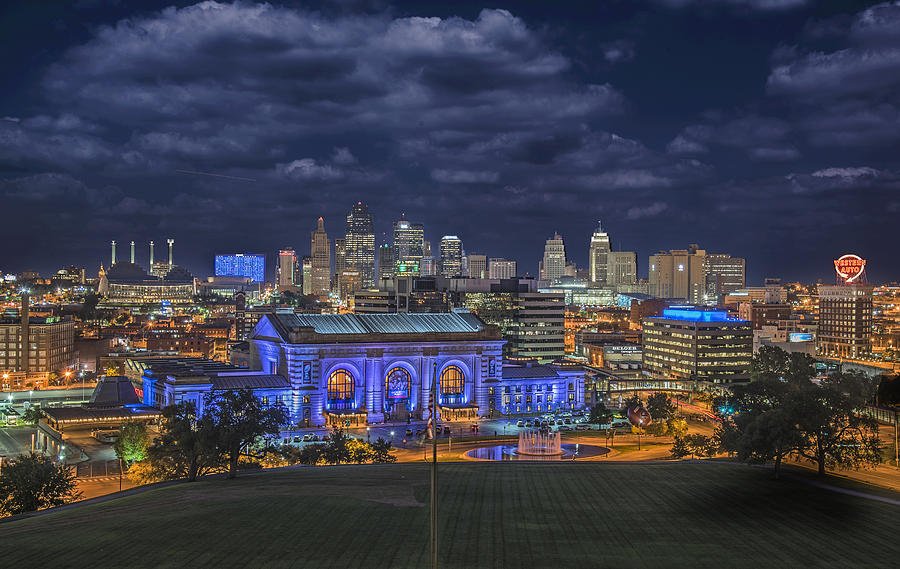 Night view of a city skyline with modern and historic buildings, illuminated with colorful lights, including a prominent train station with blue lighting and a fountain in the foreground.