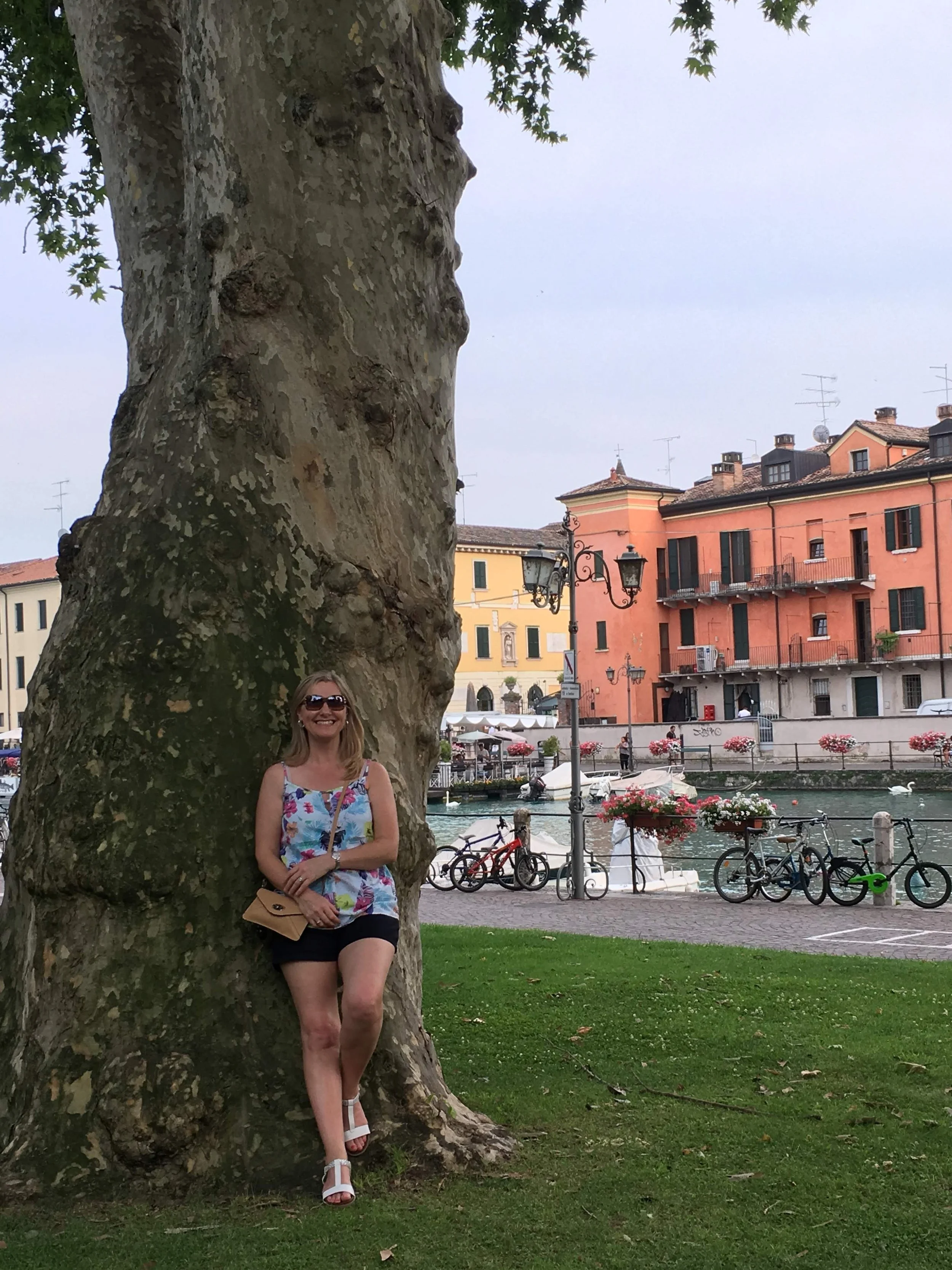 A woman in sunglasses, a floral top, and black shorts standing and leaning against a large tree with textured bark, smiling at the camera in a scenic European town with colorful buildings, bicycles, a canal, and flowers in the background.