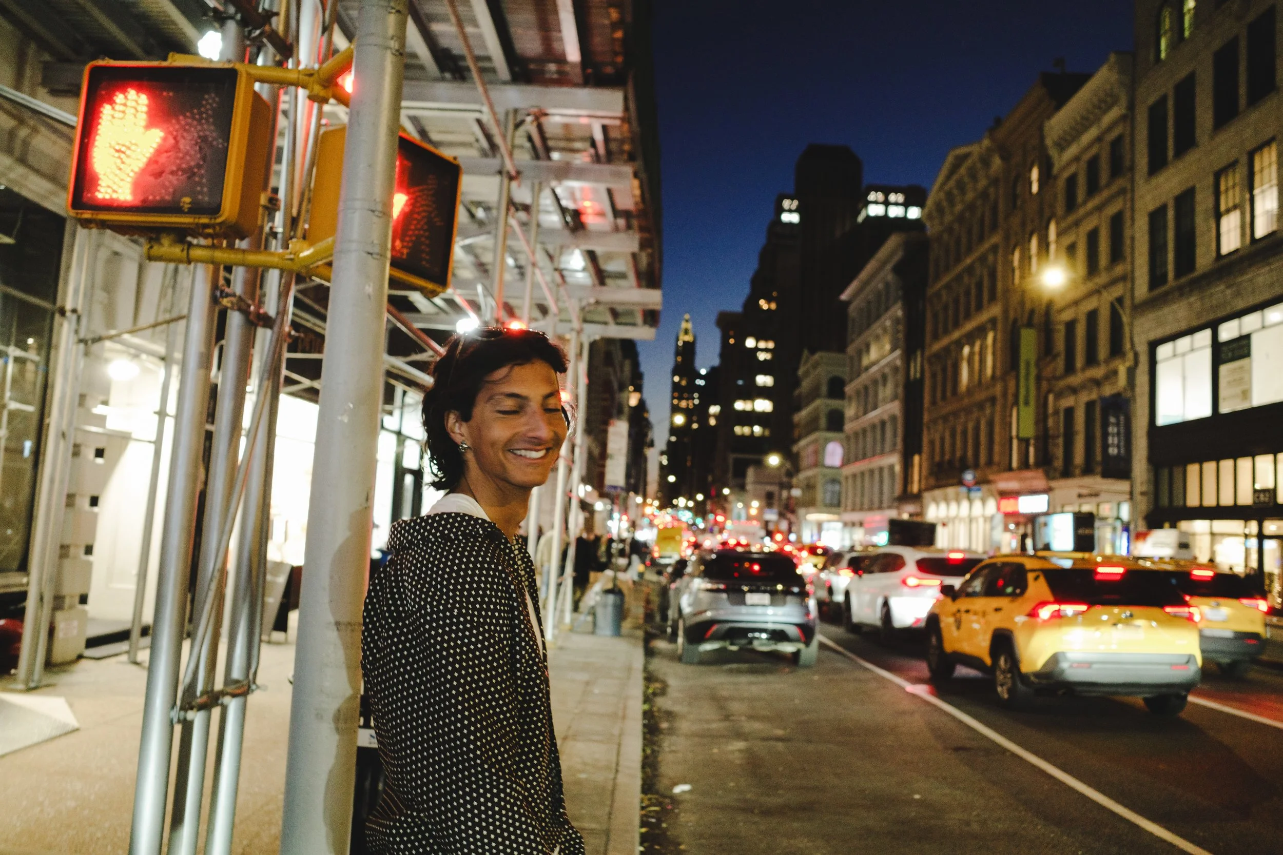 A person with short dark hair in a polka-dot coat stands on a city street at night, smiling with eyes closed. The street is busy with cars and illuminated buildings in the background. Traffic lights show a red hand signal.