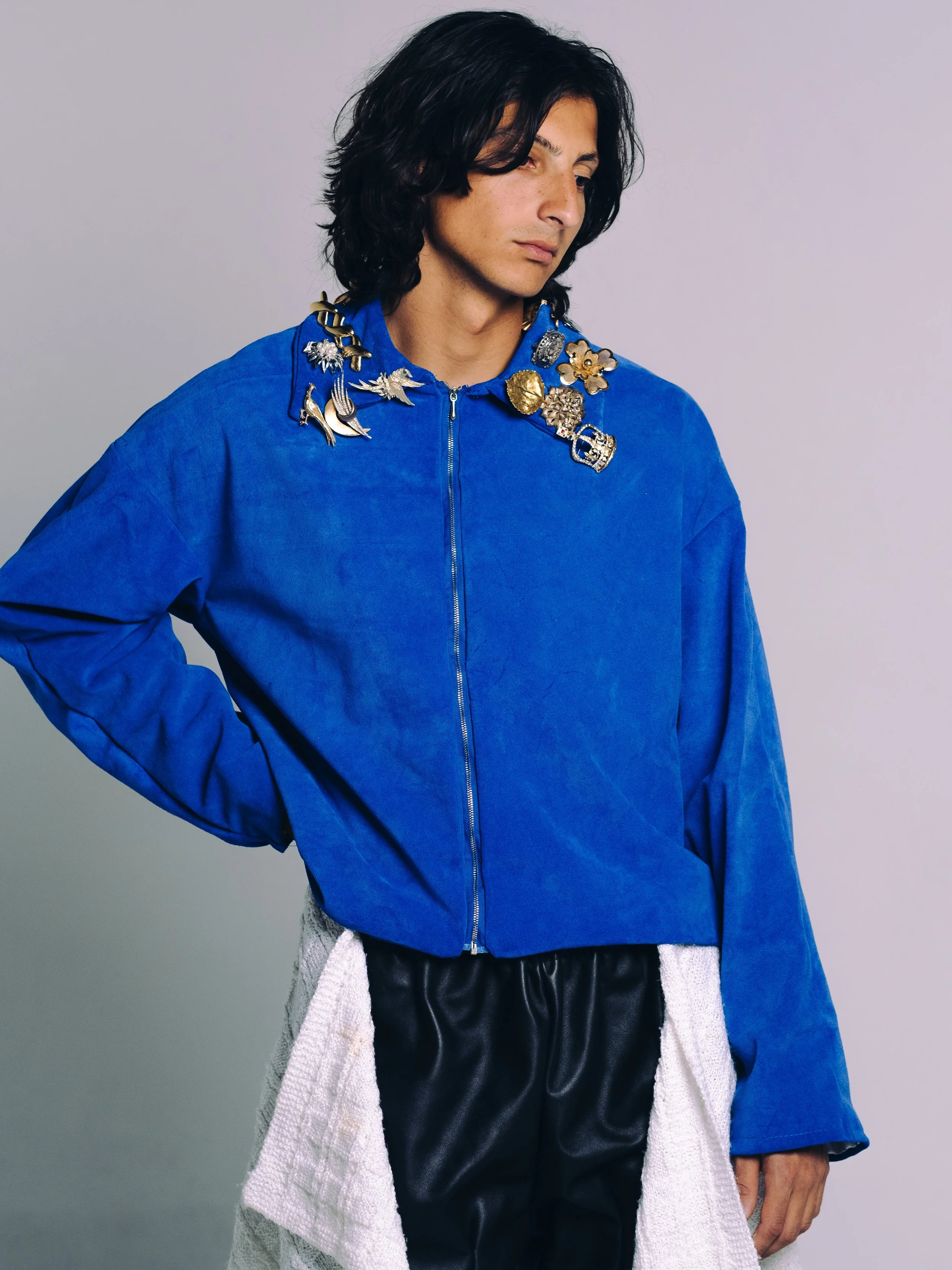 A person with long dark hair wearing a bright blue zip-up jacket with decorative pins around the collar and black leather pants with a white textured fabric hanging on the side.