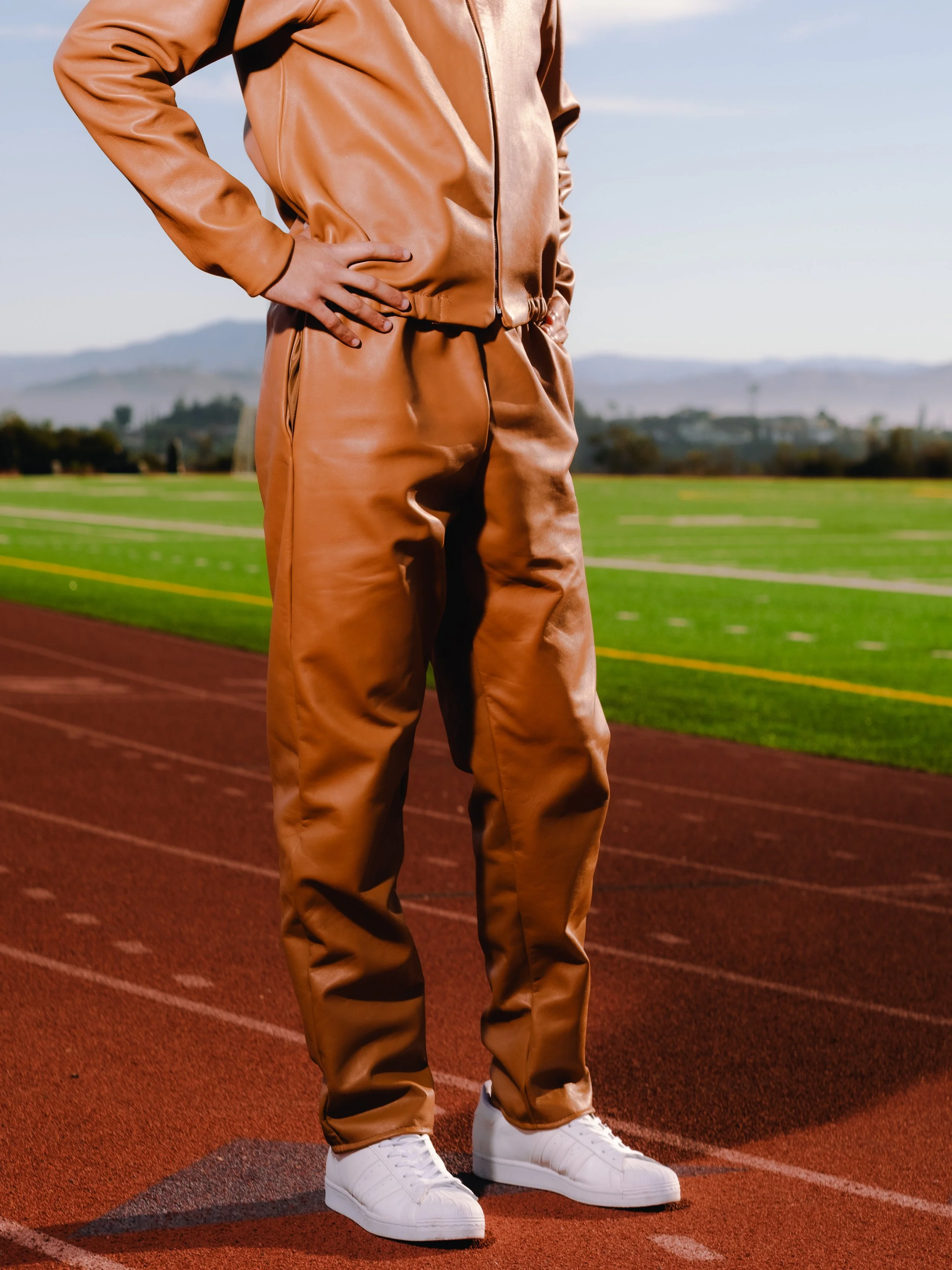 Person standing on a running track in a brown leather tracksuit and white sneakers, with a sports field and mountains in the background.