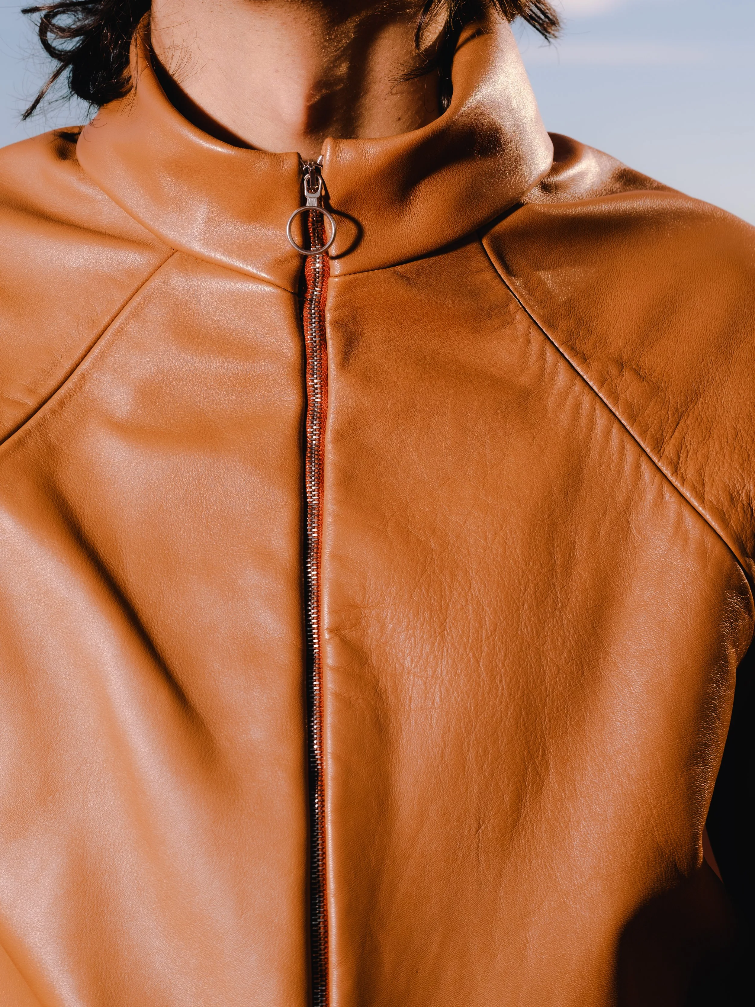 Close-up of a person wearing a tan leather jacket with a zipper up the front, the collar slightly raised, and the person outdoors.