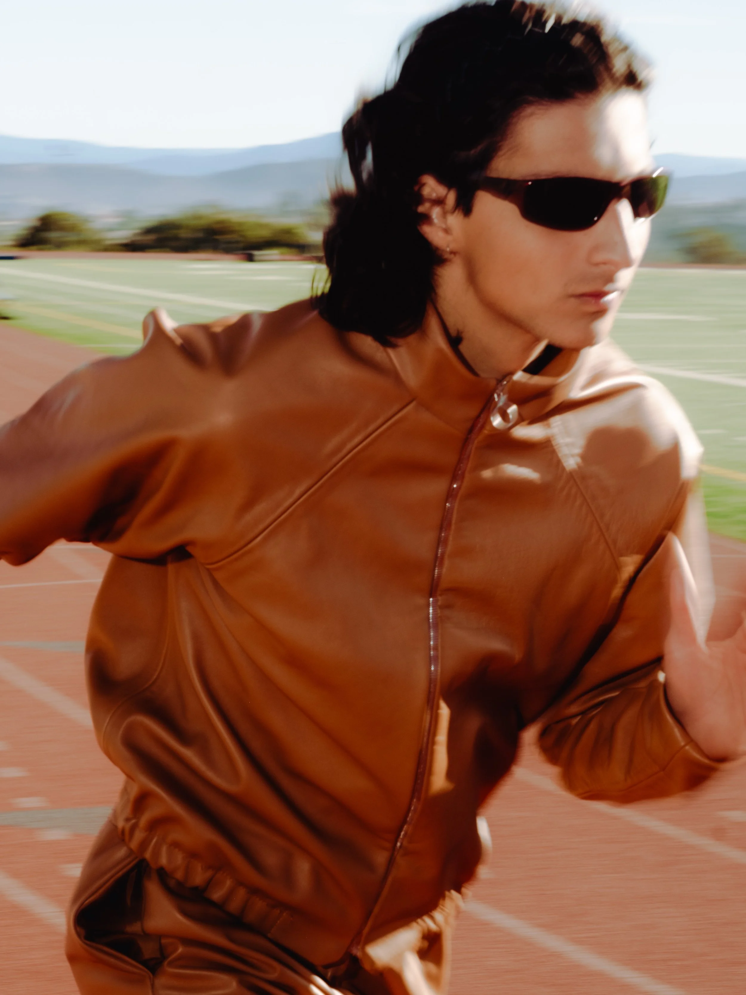A man with long dark hair wearing sunglasses and a brown leather jacket is running on a track field outdoors.