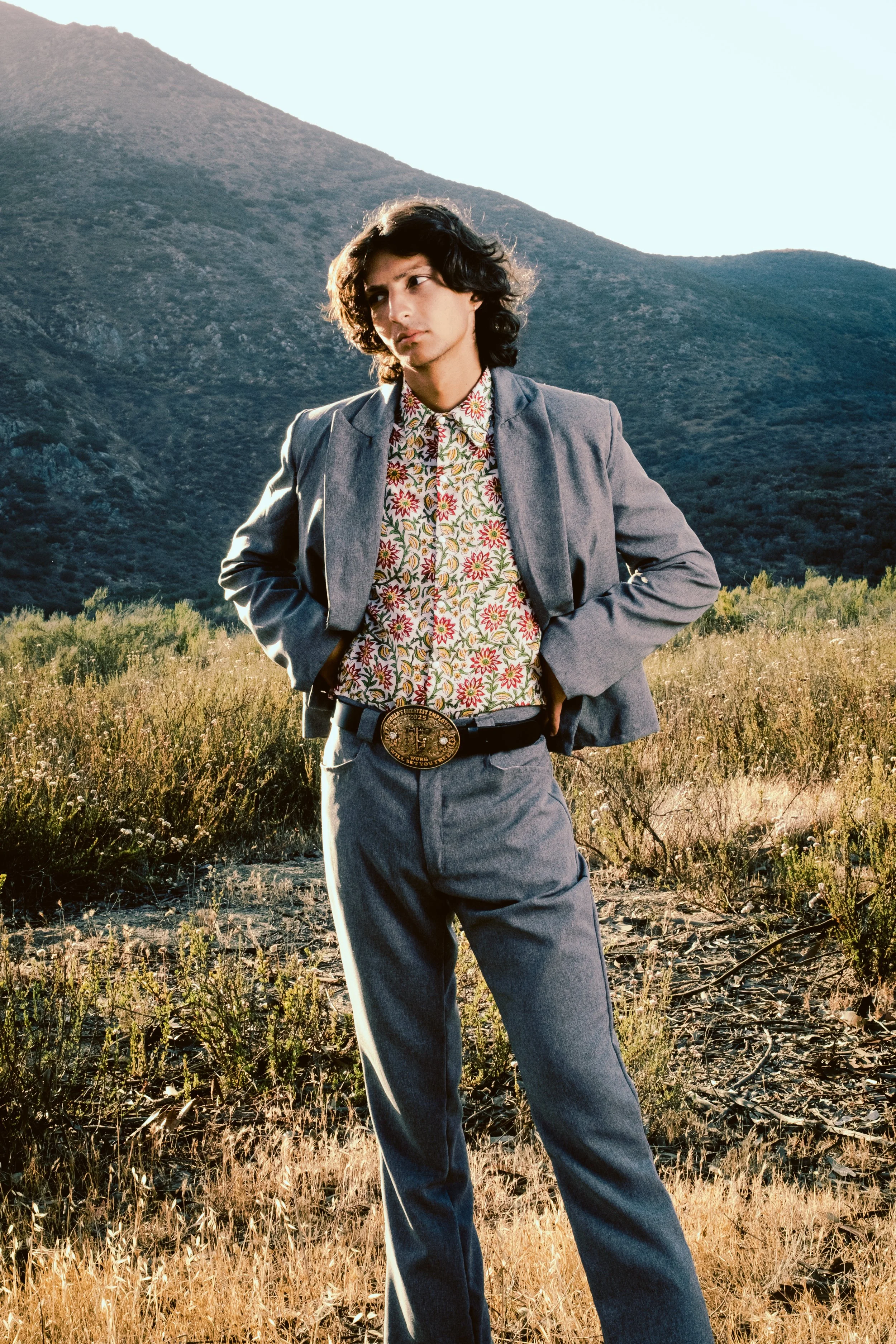 A young man with dark, curly hair standing outdoors in a field with mountains in the background. He is wearing a gray suit, floral shirt, and a large belt buckle.