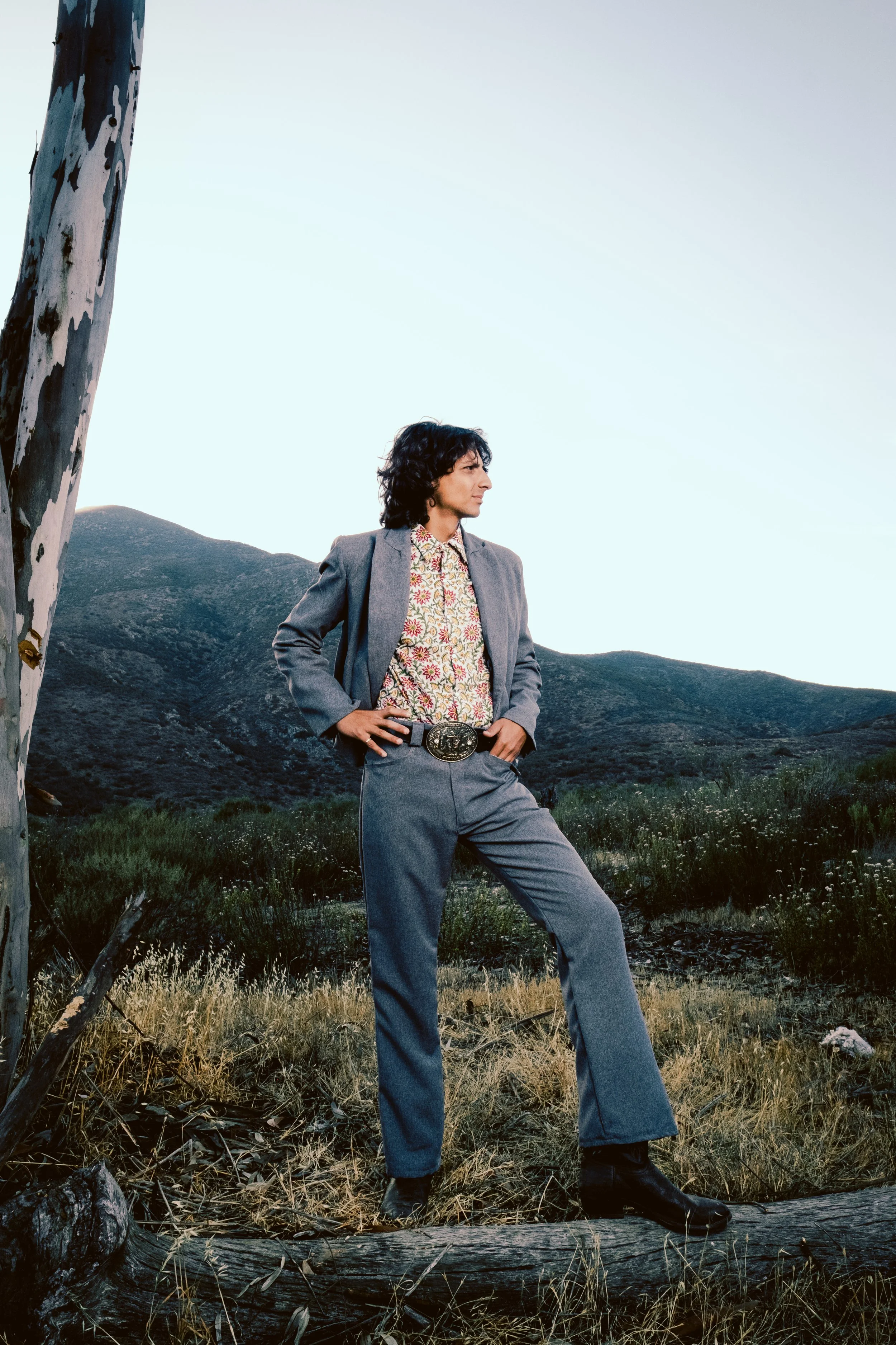 A person with dark hair wearing a gray blazer, floral shirt, and gray pants with a large belt buckle, standing outdoors in a mountainous landscape with a tree in the foreground.