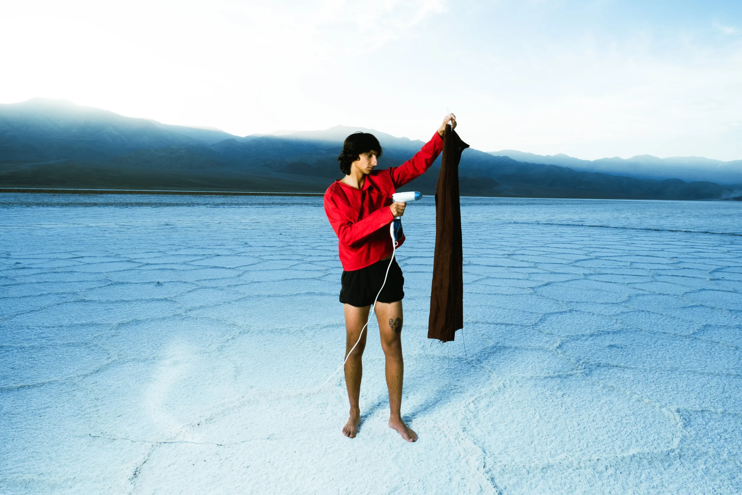 Young man in red jacket and black shorts steaming clothes with a steamer on a salt flat