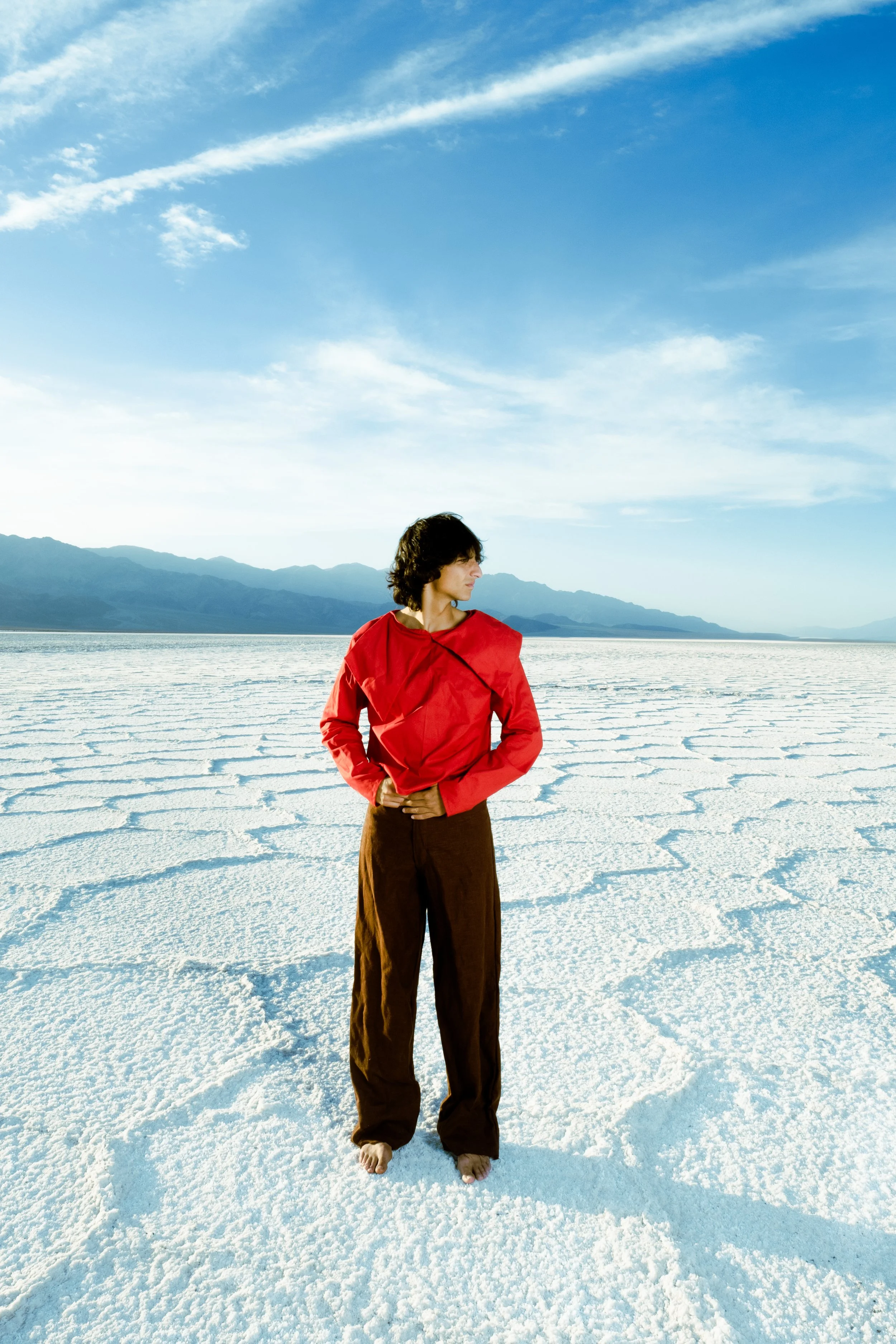 Person wearing a red jacket and brown pants standing barefoot on a salt flat with mountains in the background and a blue sky with clouds
