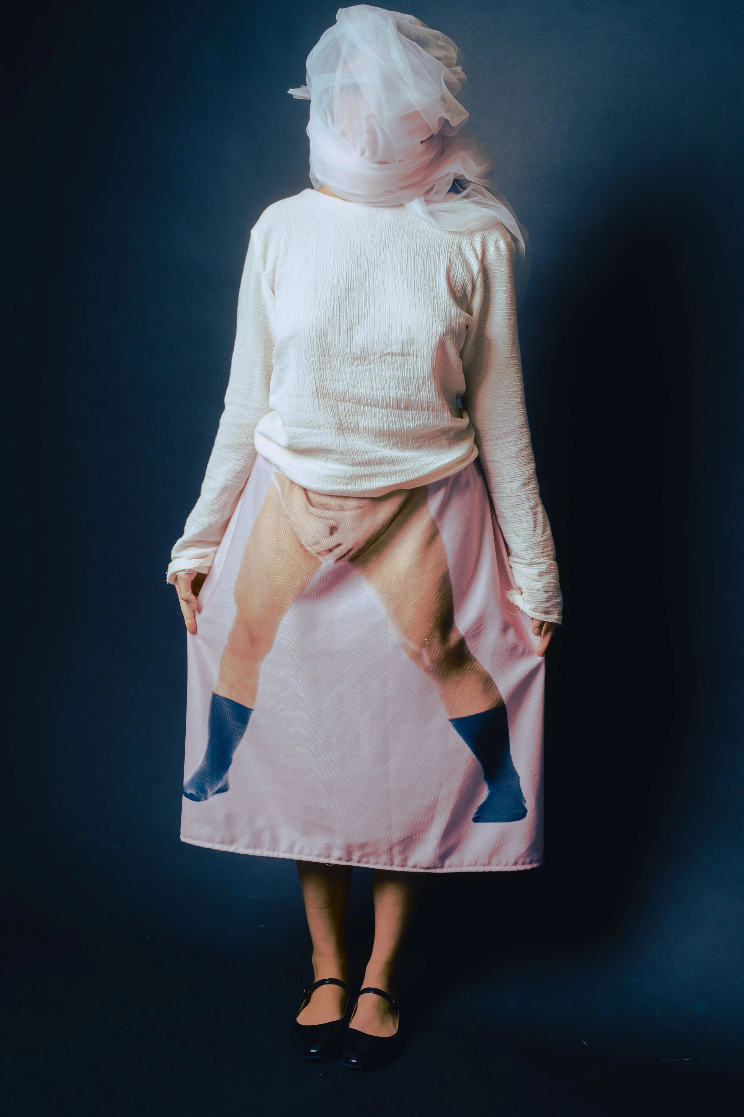 Person standing against a dark background, wearing a white long-sleeve top, a skirt with a print of a woman's legs, and black shoes, with her face covered by a white cloth.