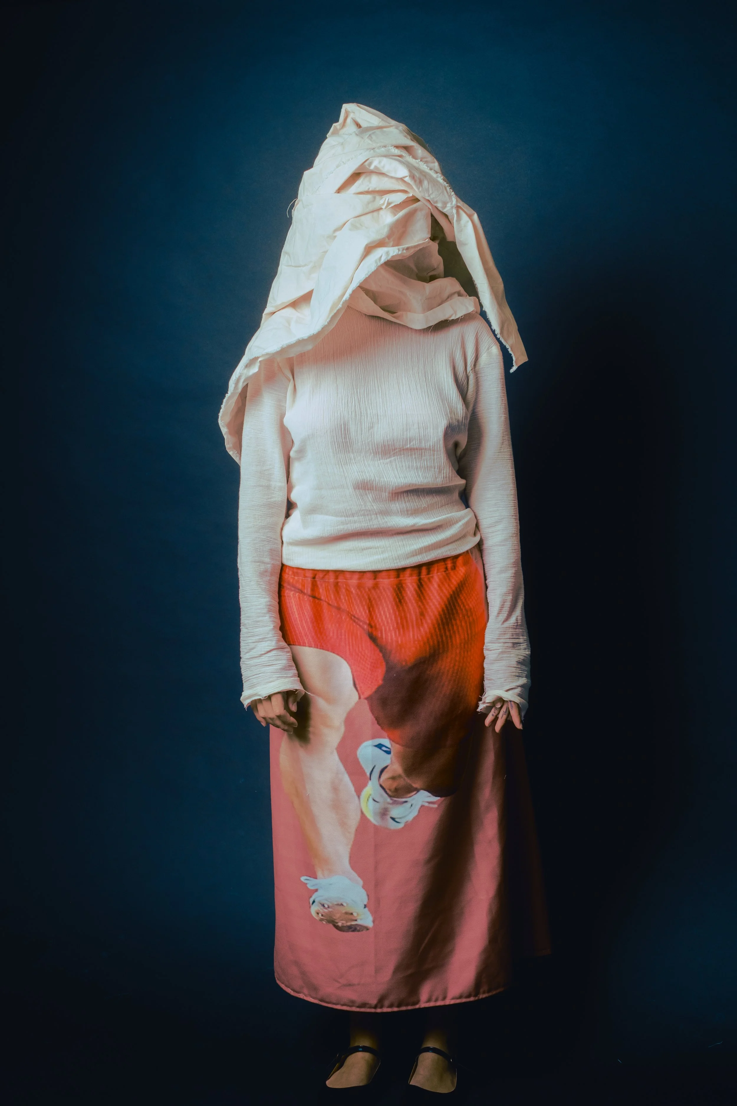 Person standing against a dark background, wearing a white long-sleeve shirt, a skirt with a large image of a person in red shorts and running shoes, and a large fabric draped over their head.