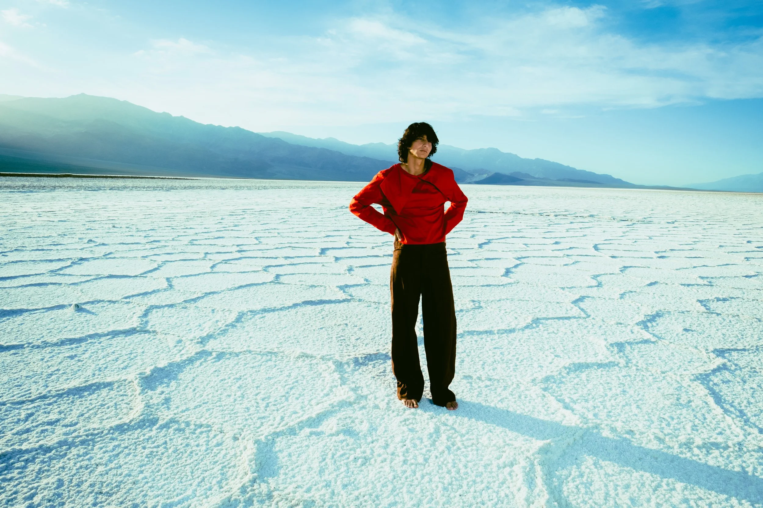Person standing on a white, cracked salt flat with mountains and a blue sky in the background, wearing a red jacket and black pants.