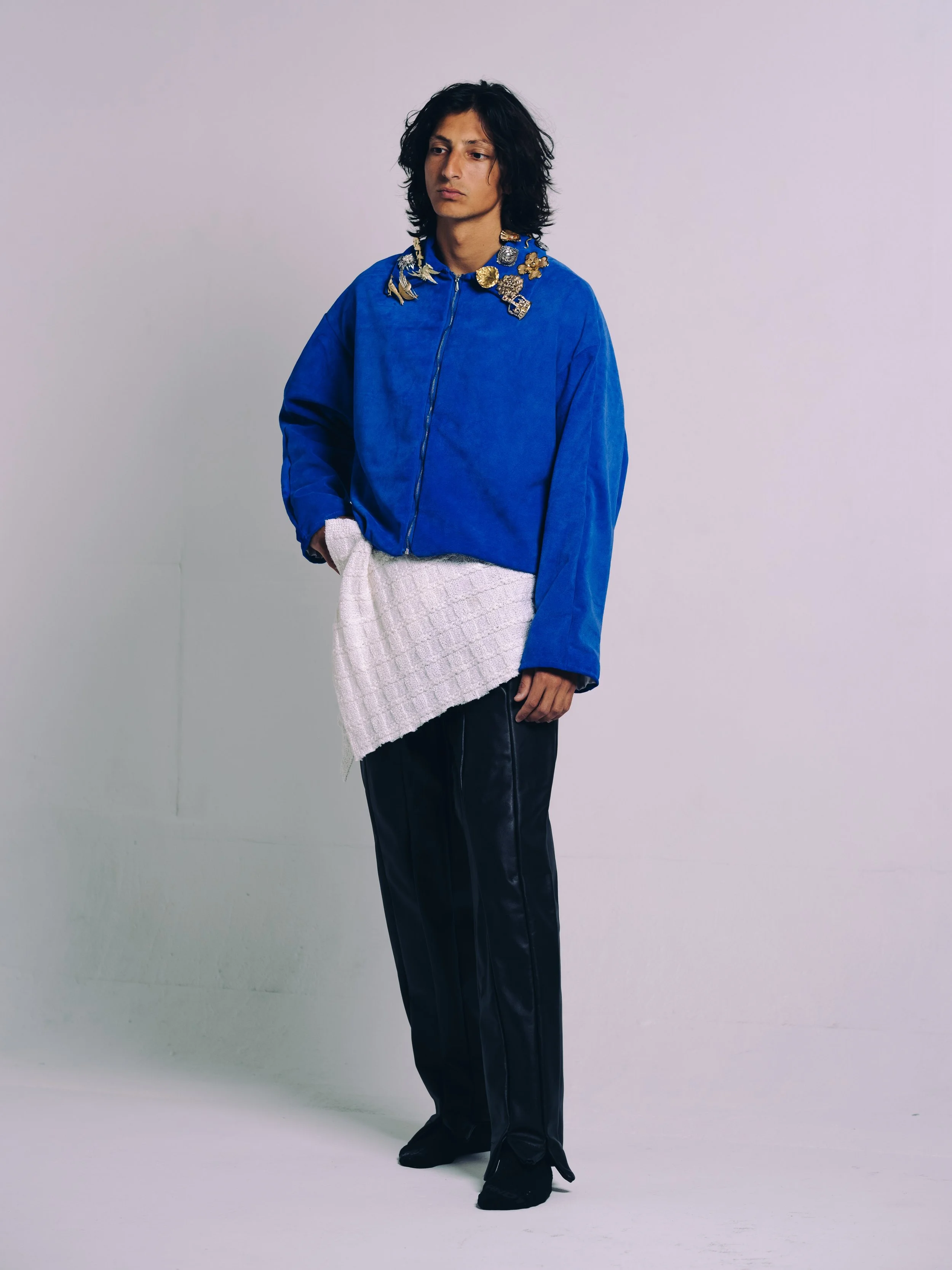 Person wearing a blue jacket with decorative pins, a white textured skirt, and black pants, standing against a plain background.