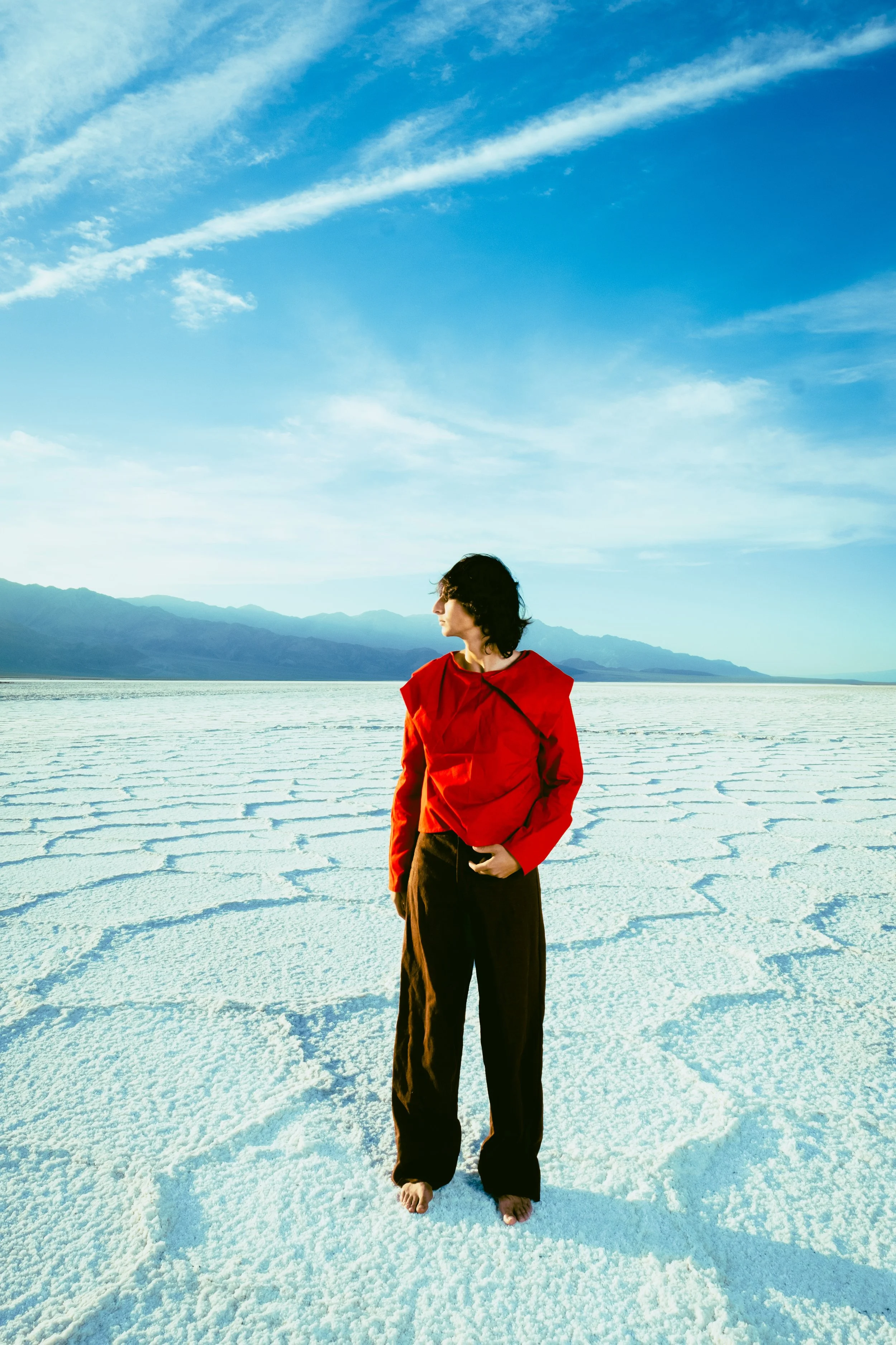 Person in red jacket and black pants standing barefoot on a white salt flat landscape with mountains in the background and blue sky with wispy clouds.