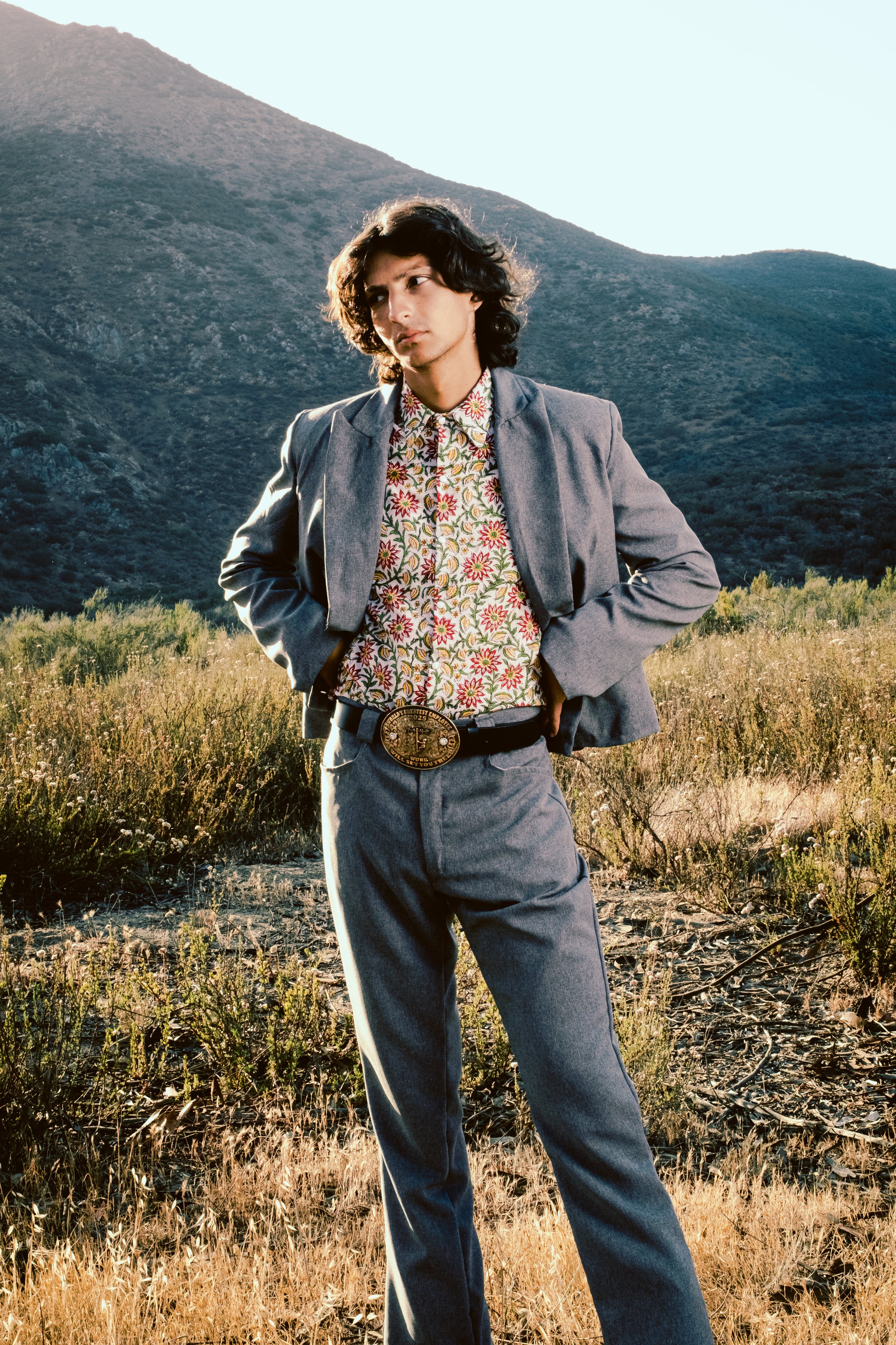 A young person standing outdoors in a grassy area with mountains in the background, wearing a gray suit, floral shirt, and large belt buckle, with hands on hips and a thoughtful expression.