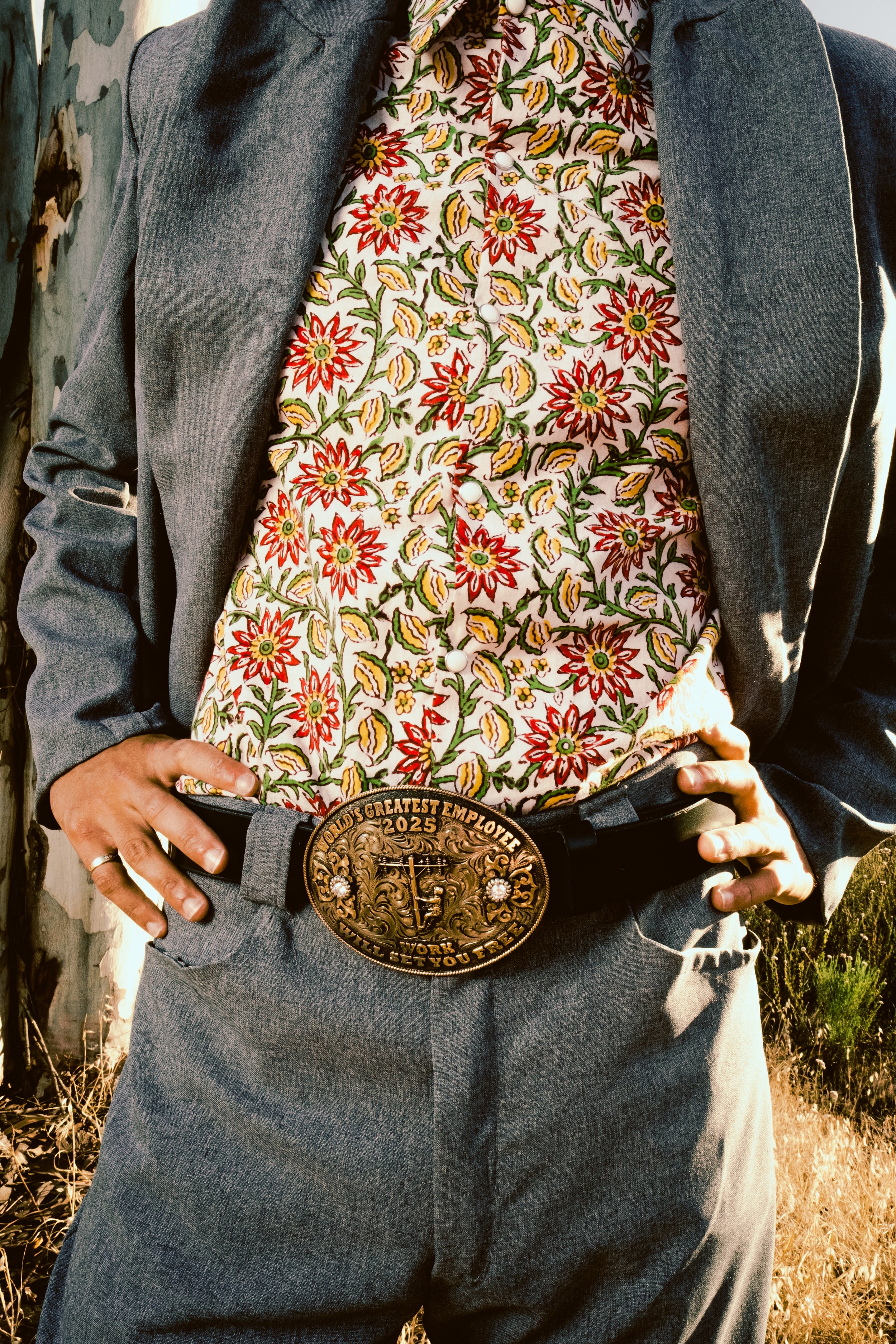 A person wearing a gray suit jacket, a colorful floral shirt, a large belt buckle that says 'World's Greatest Employee 2025', and gray pants, with hands on hips.