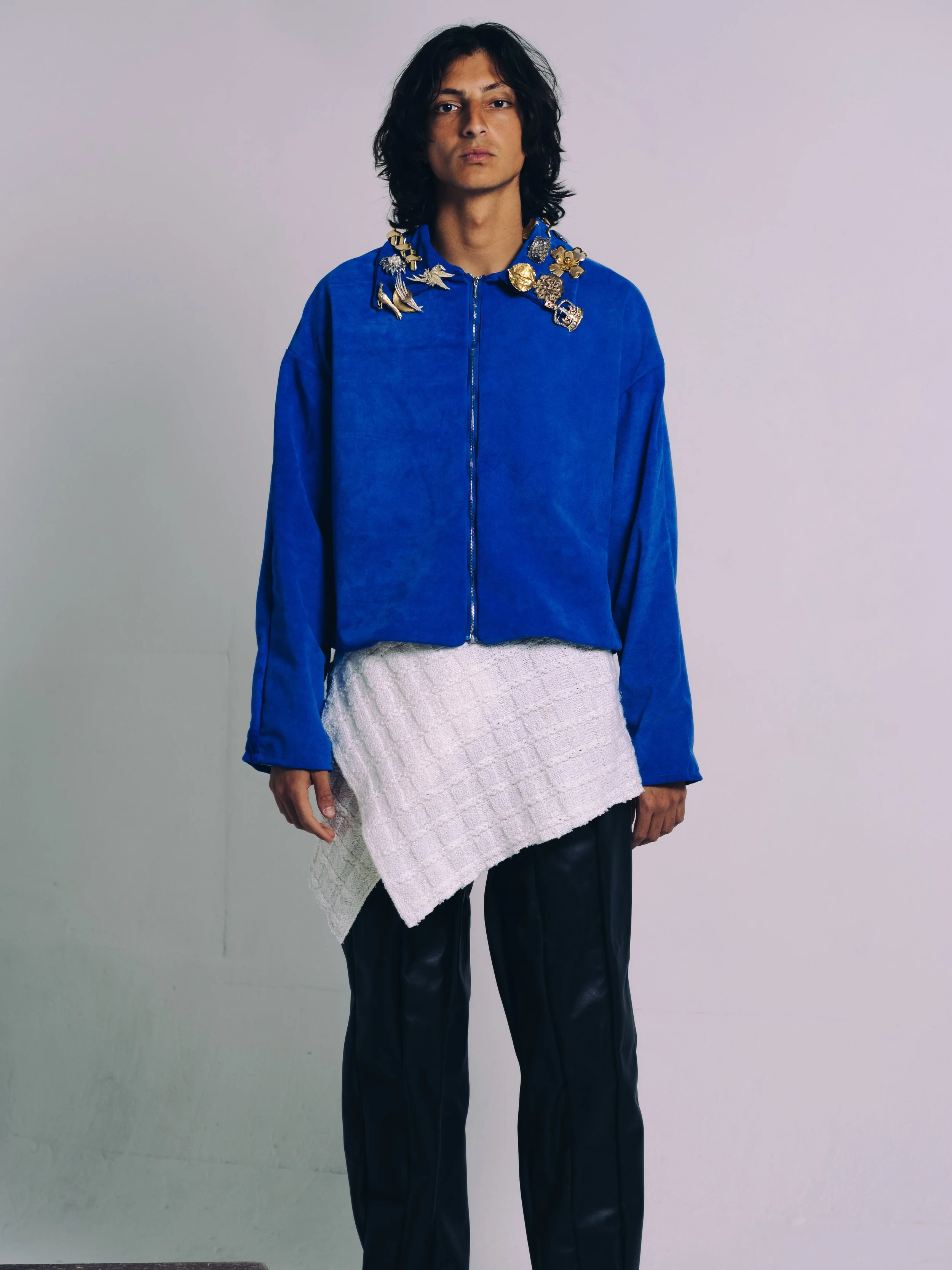 A person wearing a bright blue zip-up jacket with decorative embellishments on the collar, paired with black pants and a textured white skirt.