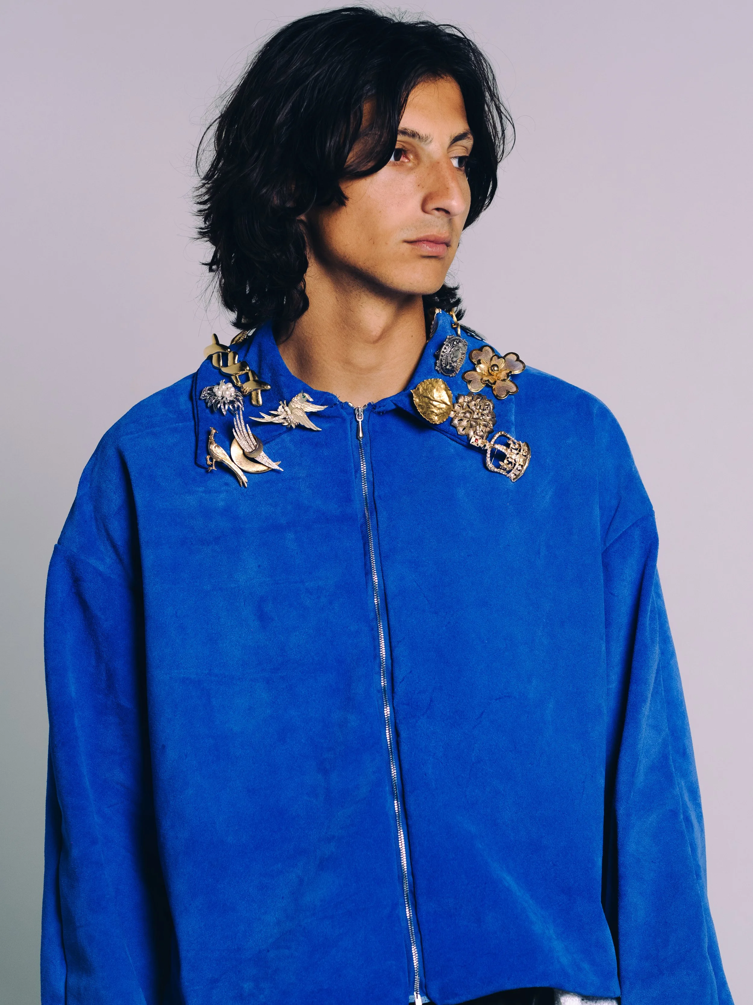 Person with dark wavy hair wearing a bright blue jacket and a statement necklace with gold and silver charms.