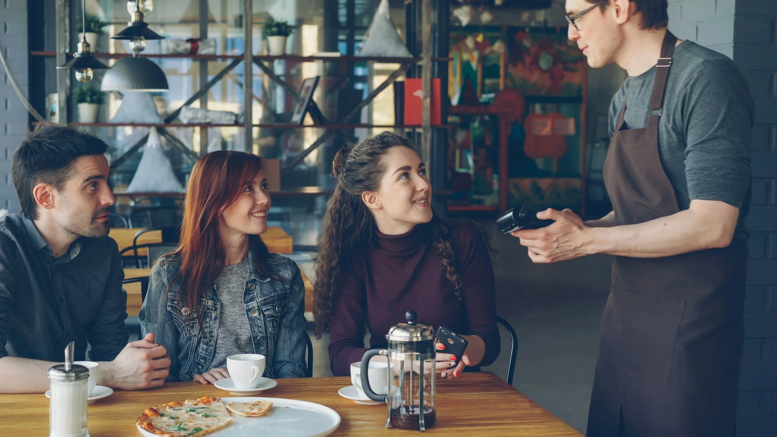 Four people sitting at a table in a cafe talking to a waiter. The table has pizza, cups, and a French press coffee maker.
