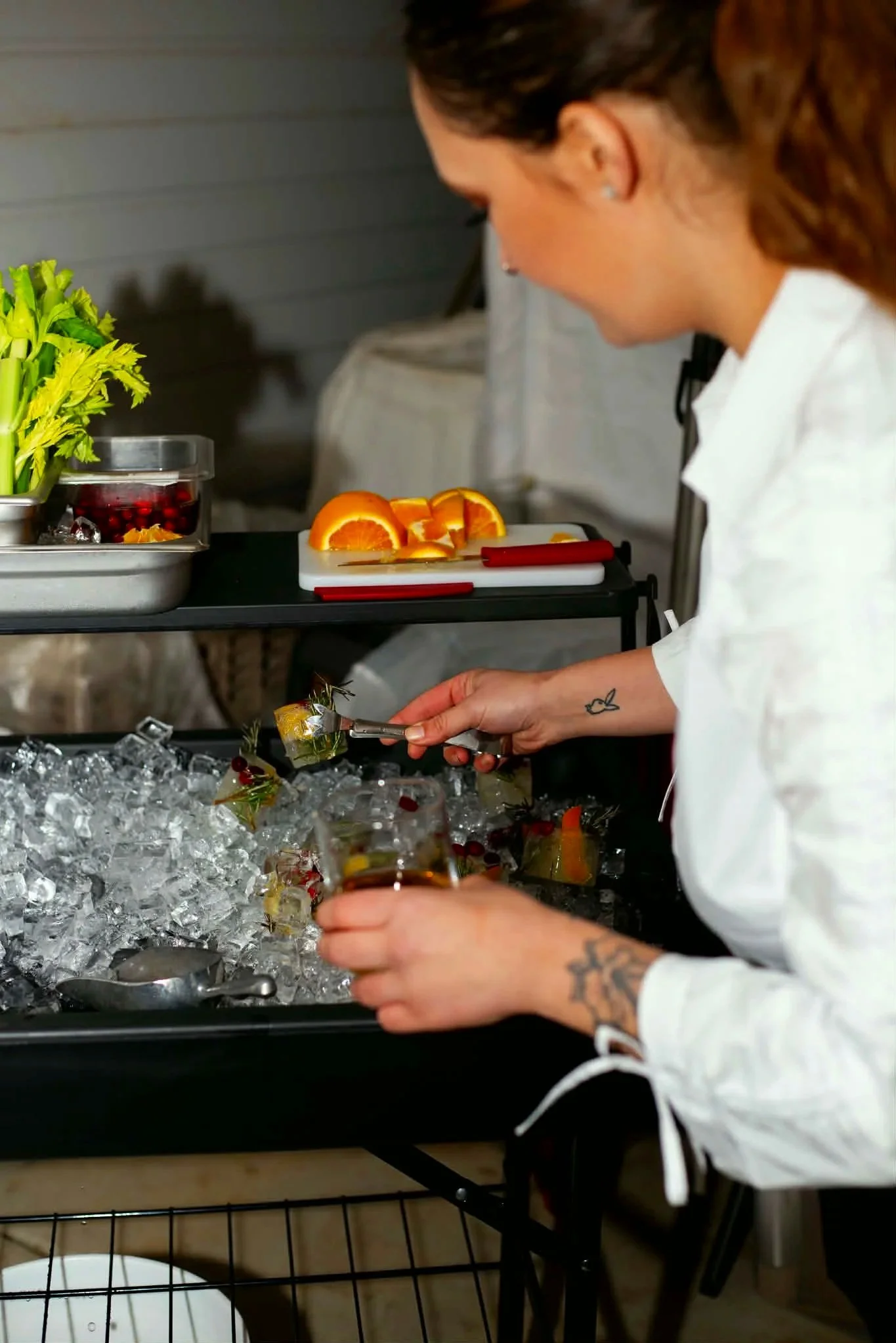 A woman with tattoos on her arms serving drinks from a tray filled with ice, with orange slices and berries, at a party or gathering.