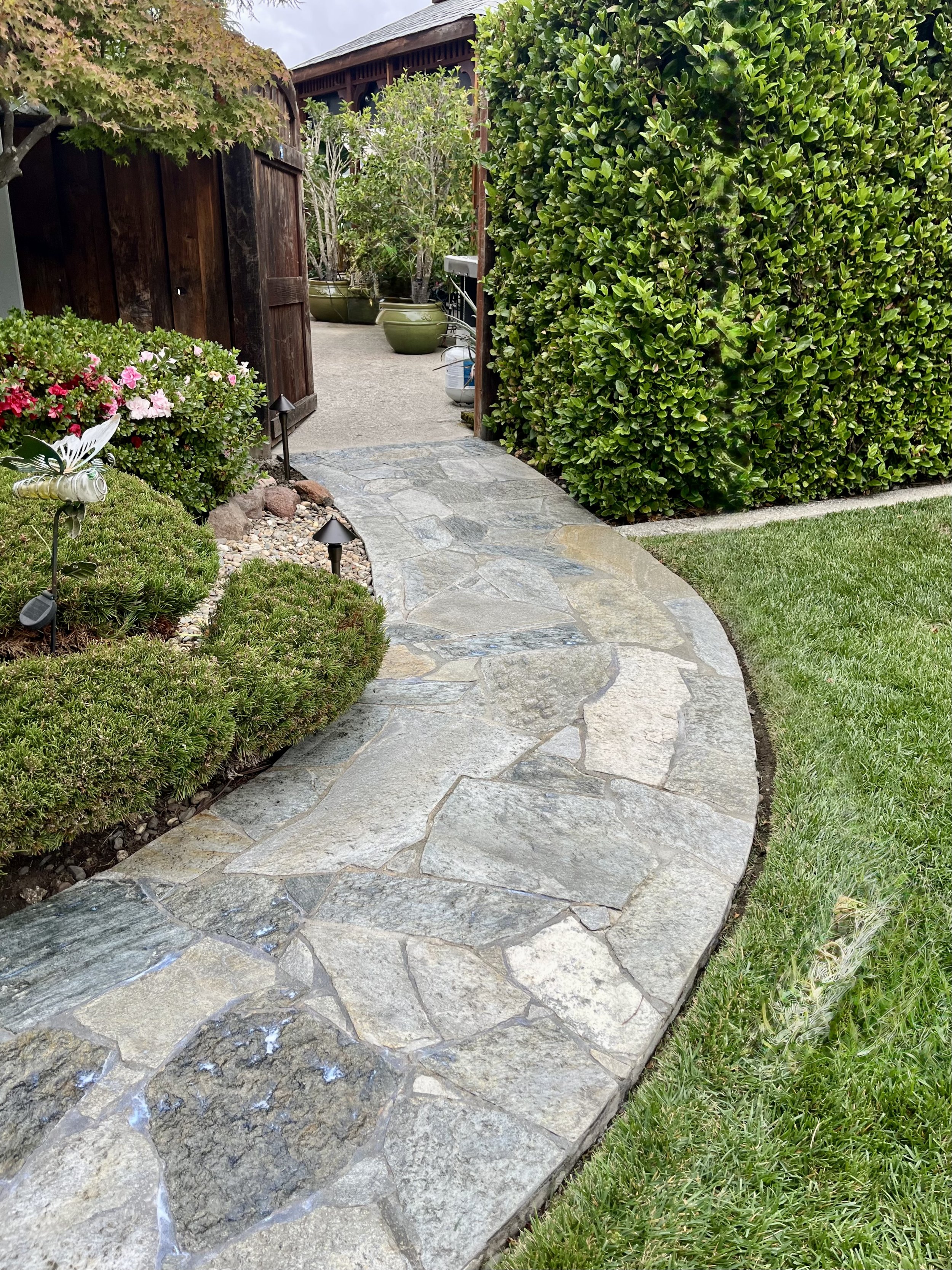 Curved stone pathway leading through a well-manicured garden with bushes, flowering plants, and large potted trees, bordered by grass and a wooden fence.