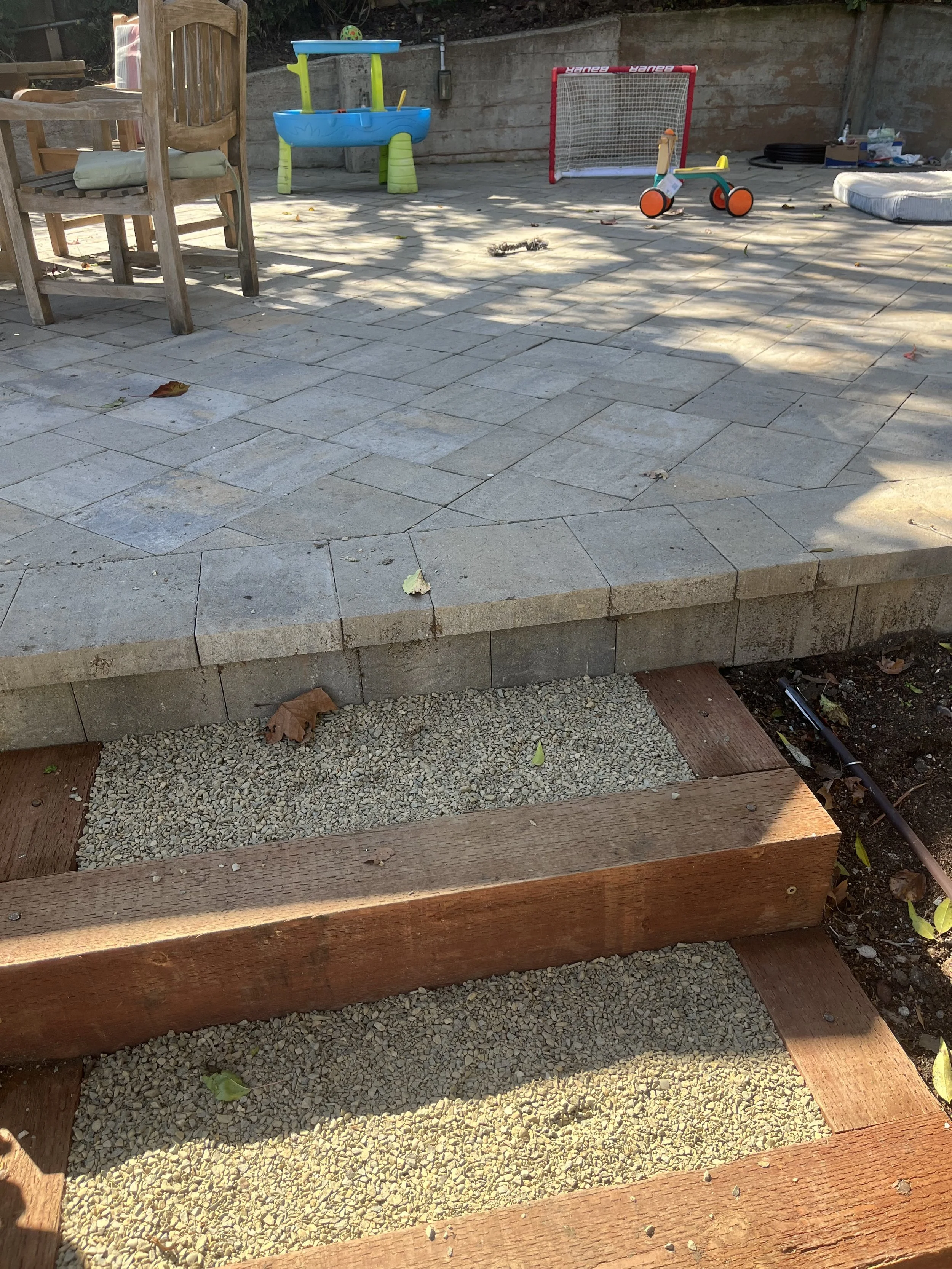 Backyard patio with wooden steps, stone tiles, children's toys, including a plastic sandbox, a small soccer goal, and a tricycle, with scattered leaves and garden tools.