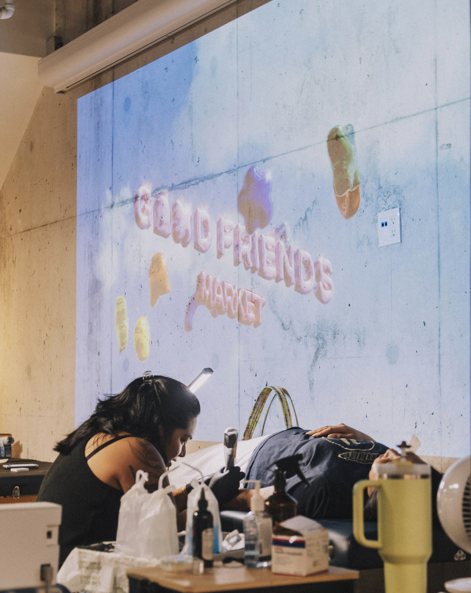 A person lying on a medical examination table with their head turned to the side, while another person is sitting nearby, possibly administering an injection. There are various medical supplies on the table in front of them. A large screen shows a colorful digital display with the words 'GRATITUDE, LOVE, HAPPINESS' in pink, along with images of brightly colored candies or decorations.