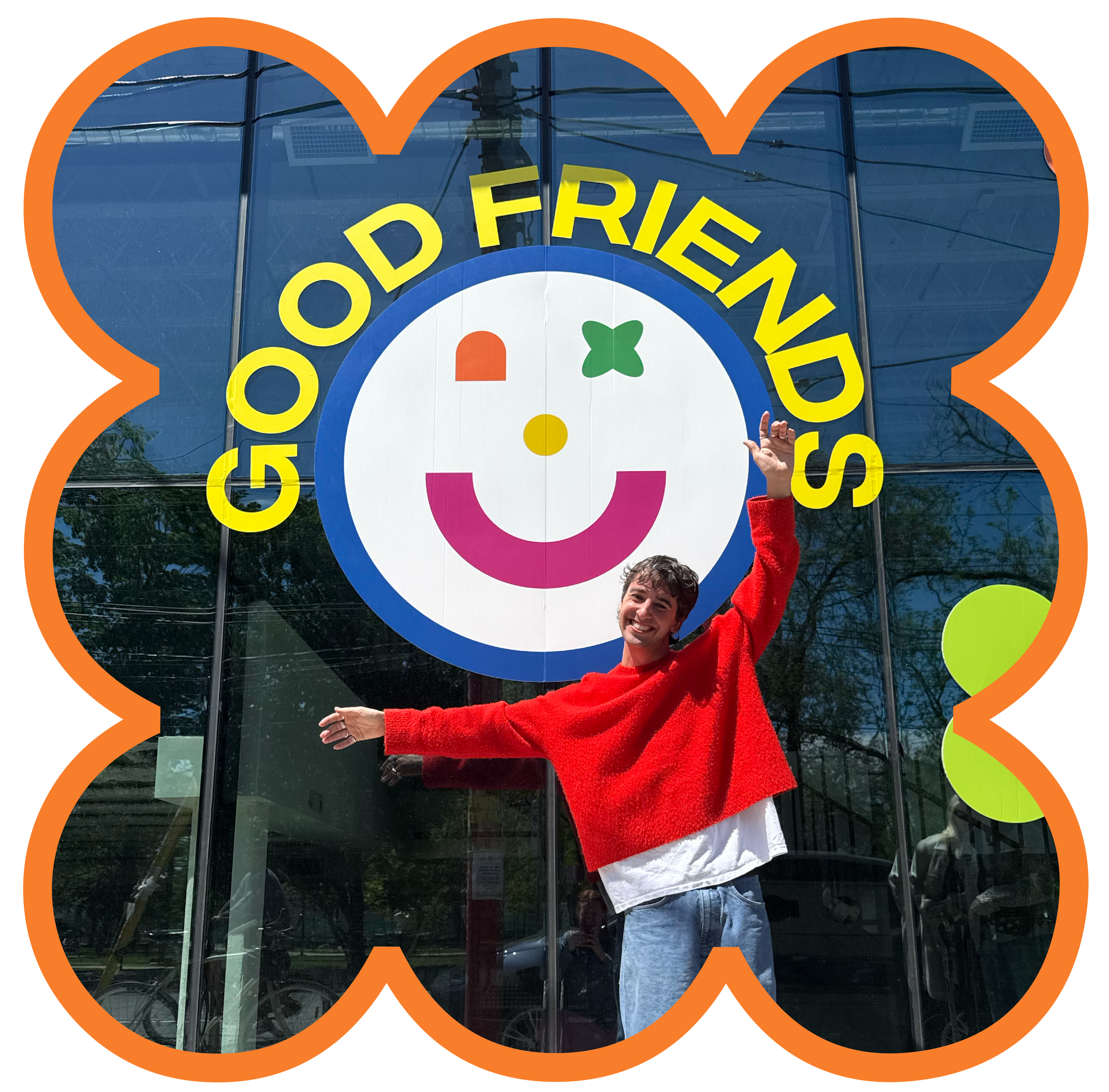 A person in a red sweater and jeans standing outside a building with glass windows, smiling and raising one arm. Behind them is a logo with a smiling face with colorful features and the words "Good Friends" in yellow. The photo is framed with an orange flower-shaped border.