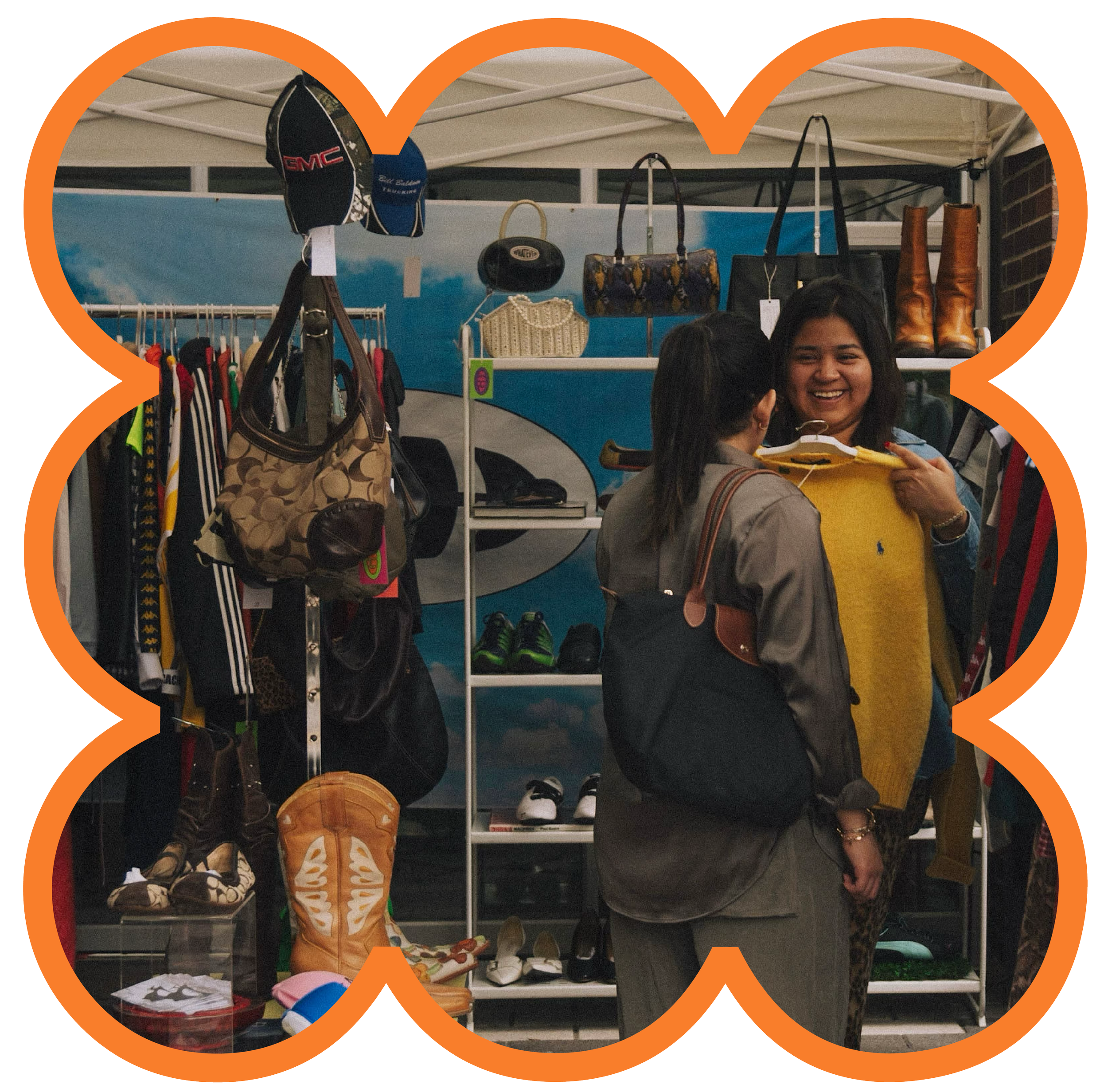 Two women shopping at a clothing and accessories stall, one woman is smiling and holding a yellow sweater, the other is facing her, both are carrying handbags.