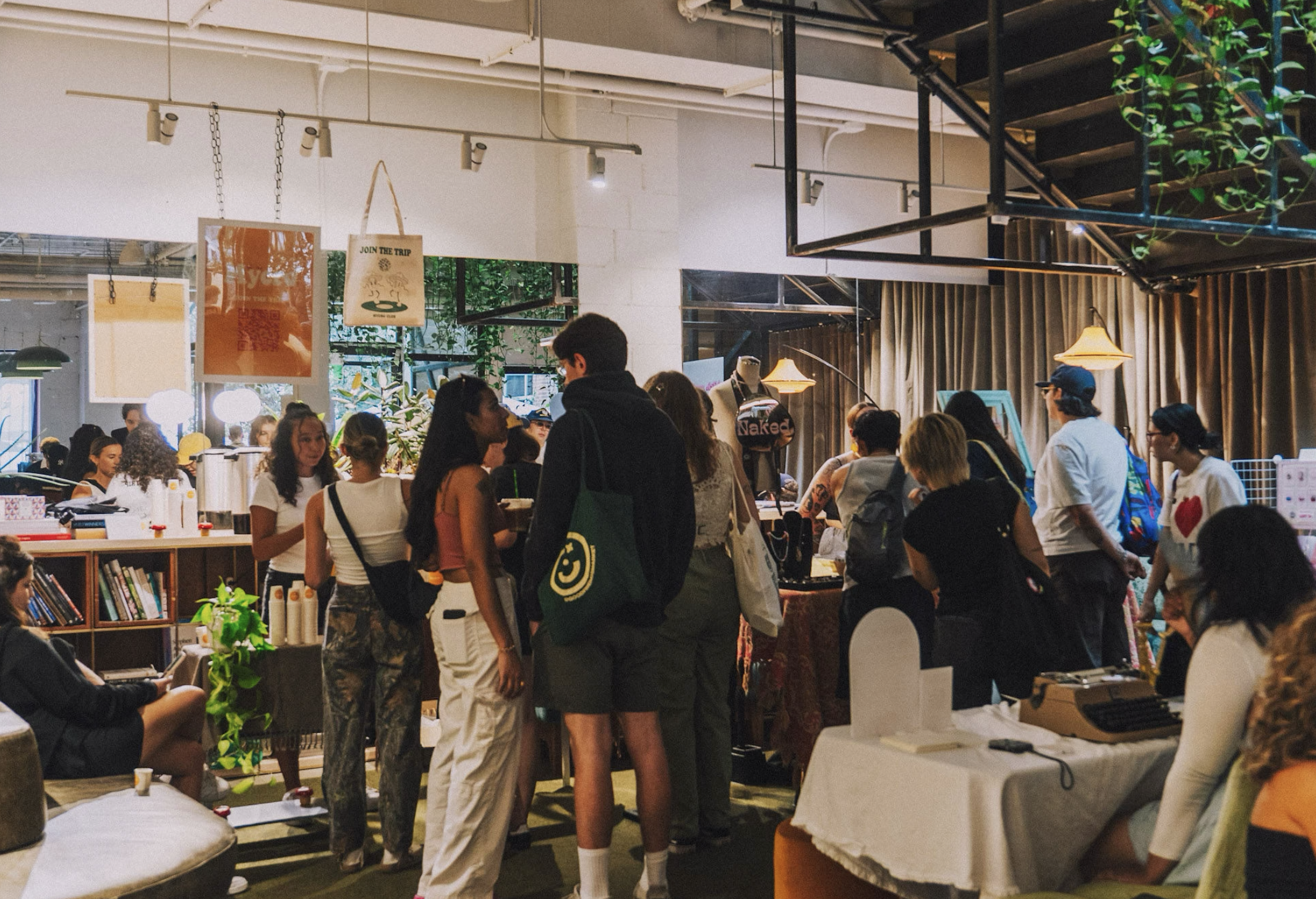 People shopping and browsing in an indoor market or boutique with various displays, bookshelves, and decorative plants.