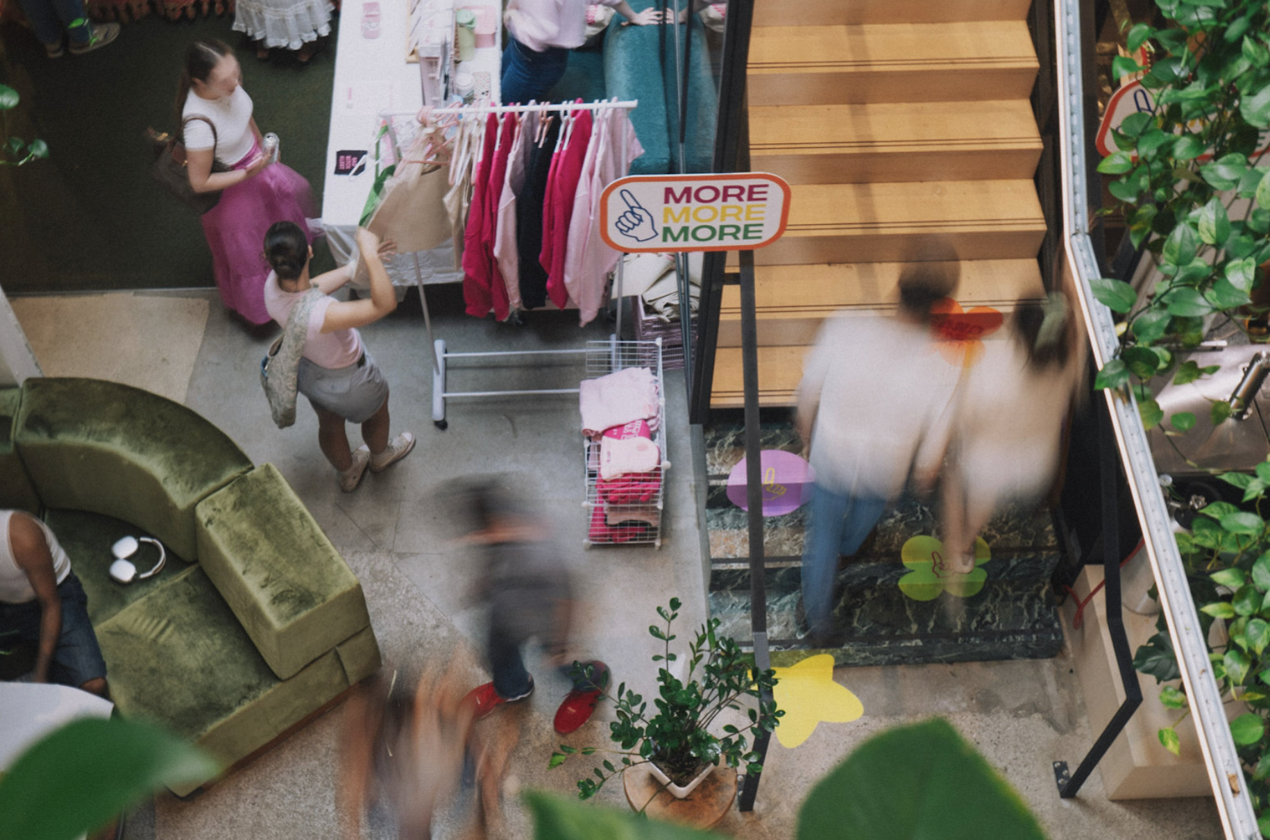 View from above of people shopping in a retail store with clothing racks, a staircase, and a seating area, with a sign saying 'More More More'.