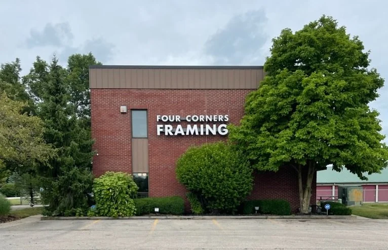 A brick building with a sign reading "Four Corners Framing," surrounded by trees and bushes, and a parking lot in the foreground.