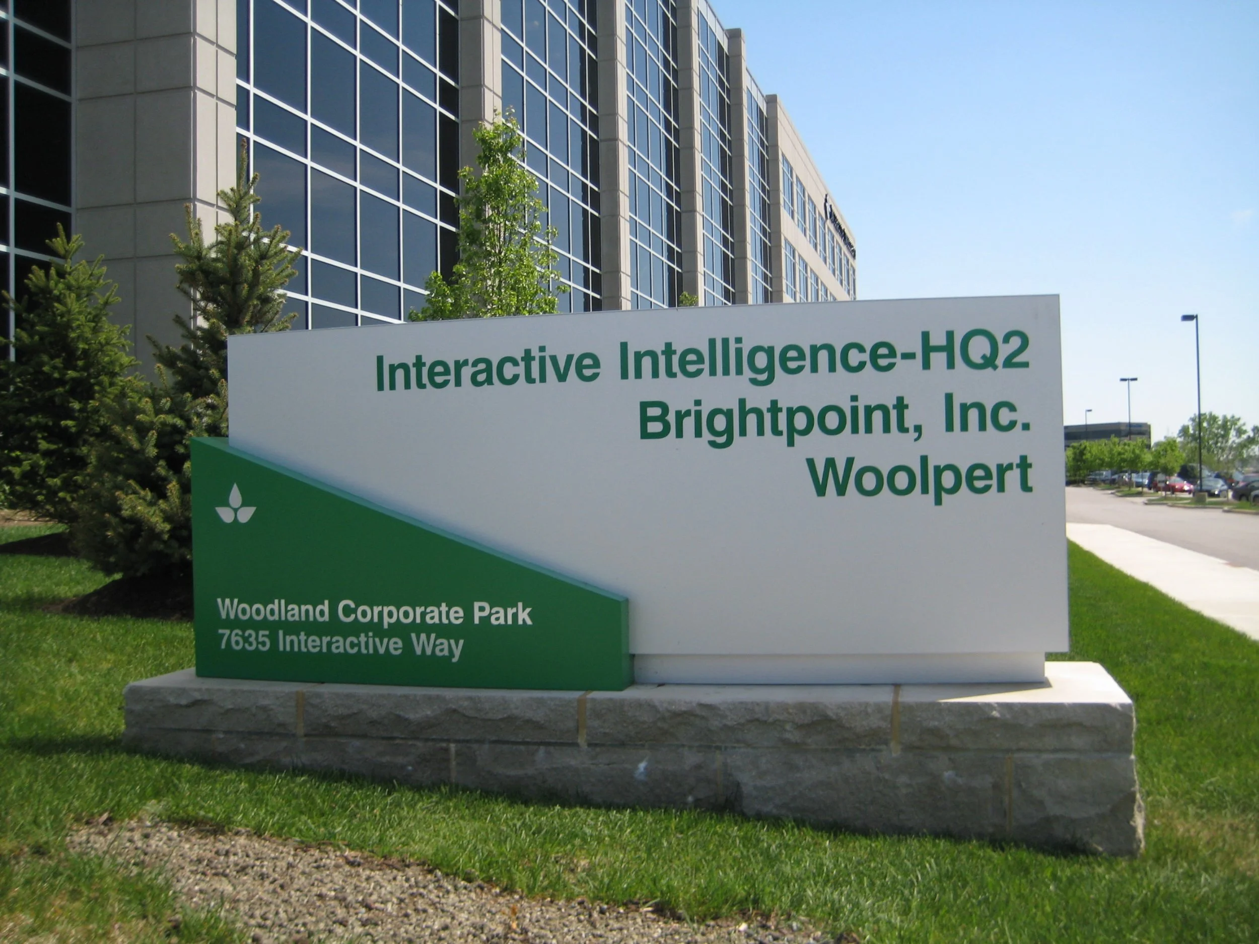 Sign for Woodland Corporate Park at 7635 Interactive Way, showing tenants Interactive Intelligence-HQ2, Brightpoint Inc., and Woolpert, in front of a modern glass office building.