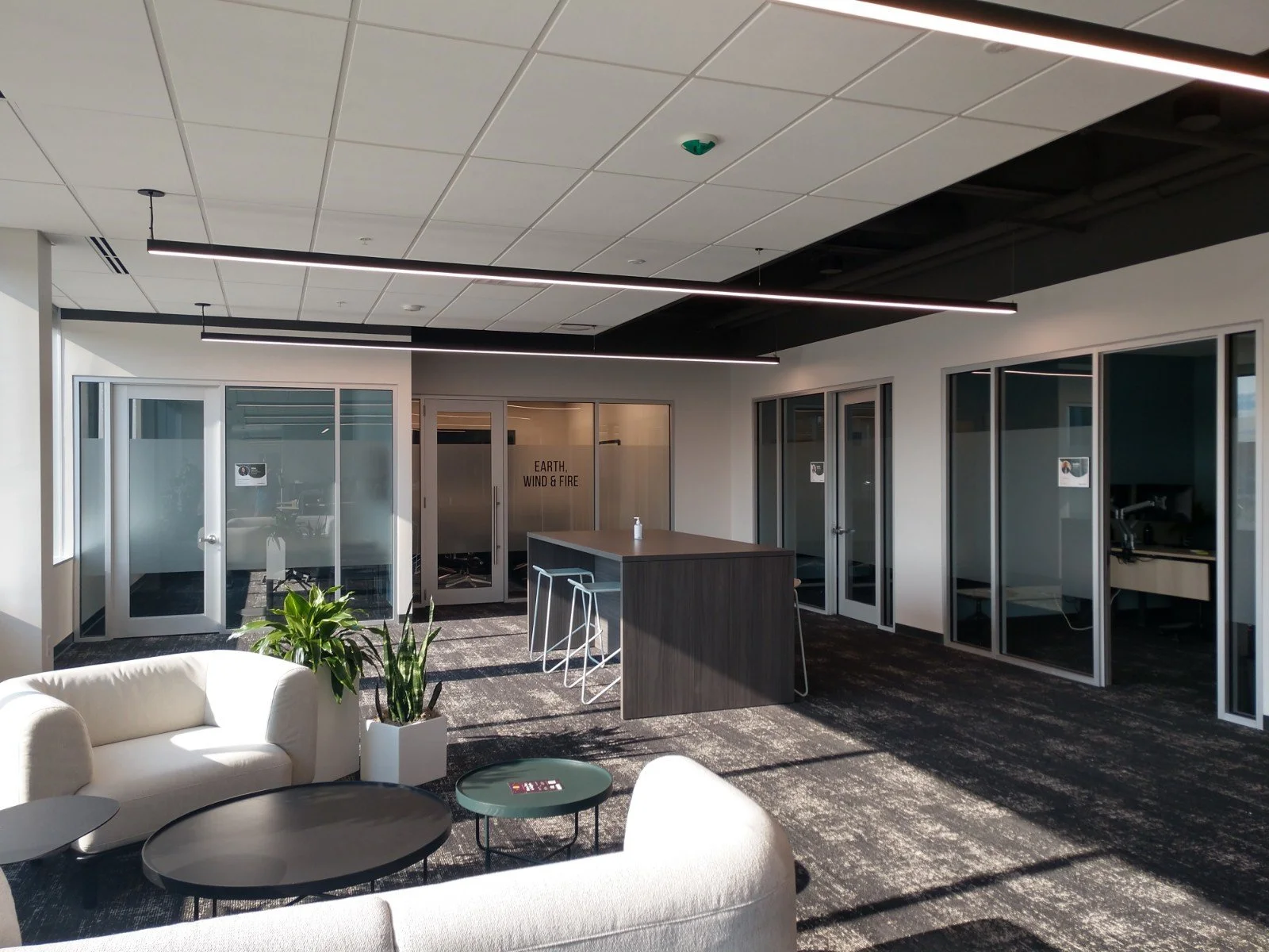 Modern office lounge with white sofas, potted plants, round tables, a kitchen island with bar stools, and glass-walled conference rooms with frosted and clear glass doors.
