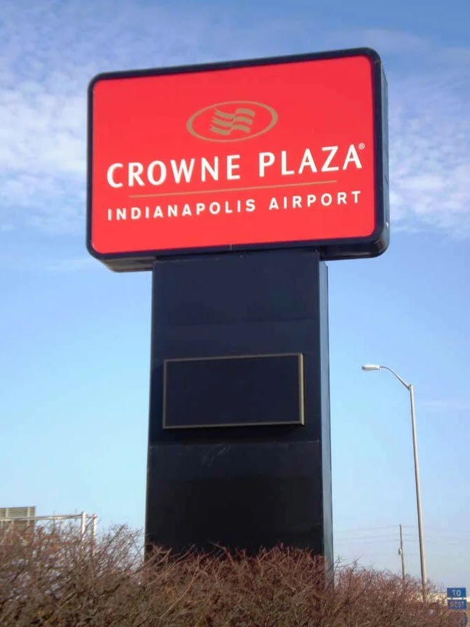 Sign for Crowne Plaza hotel at Indianapolis Airport. Red background with white text and logo, black base, with a streetlamp and cloudy sky in the background.