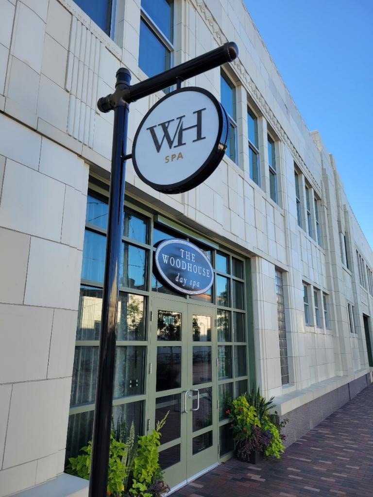 Street view of a modern building entrance with a round sign reading 'WH SPA' hanging from a black pole, and a window sign for 'The Woodhouse Day Spa' above the door, with potted plants near the door.