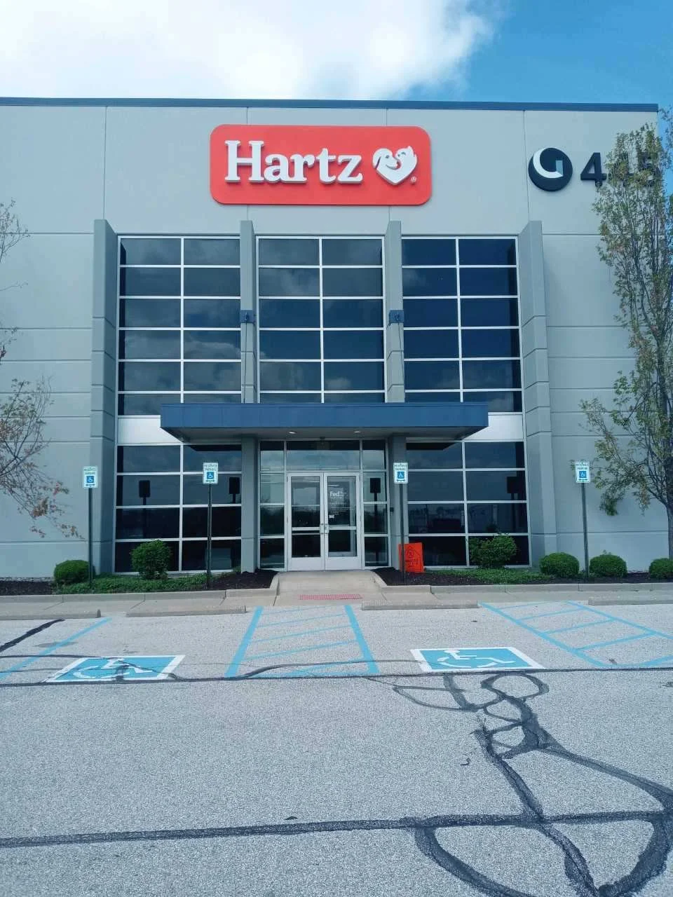 Front view of an office building with a 'Hartz' sign and a logo with two smiling faces on top. The building has large glass windows and a parking lot with designated handicapped parking spaces in front.