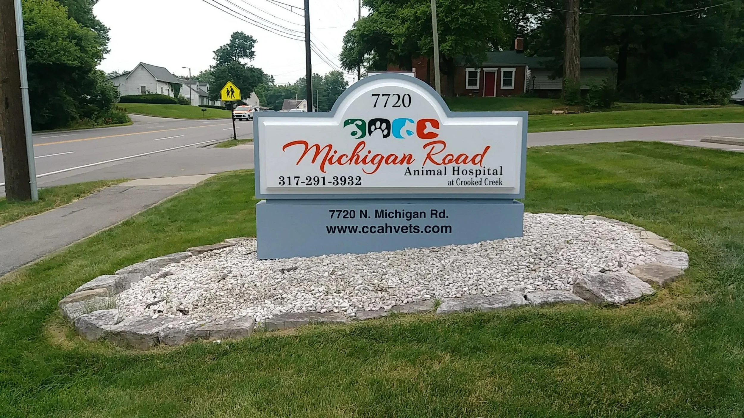 Sign for Michigan Road Animal Hospital at 7720 North Michigan Road, featuring logo and contact information, surrounded by grass and small stones, with a street and residential area in the background.