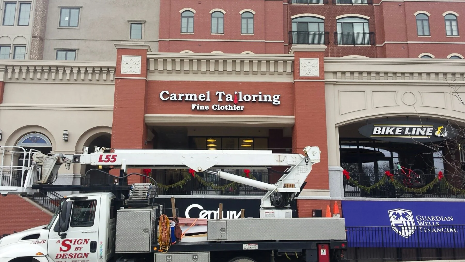 A construction vehicle parked in front of a strip mall with stores named Carmel Tailoring and Bike Line, decorated with holiday garlands and red bows.